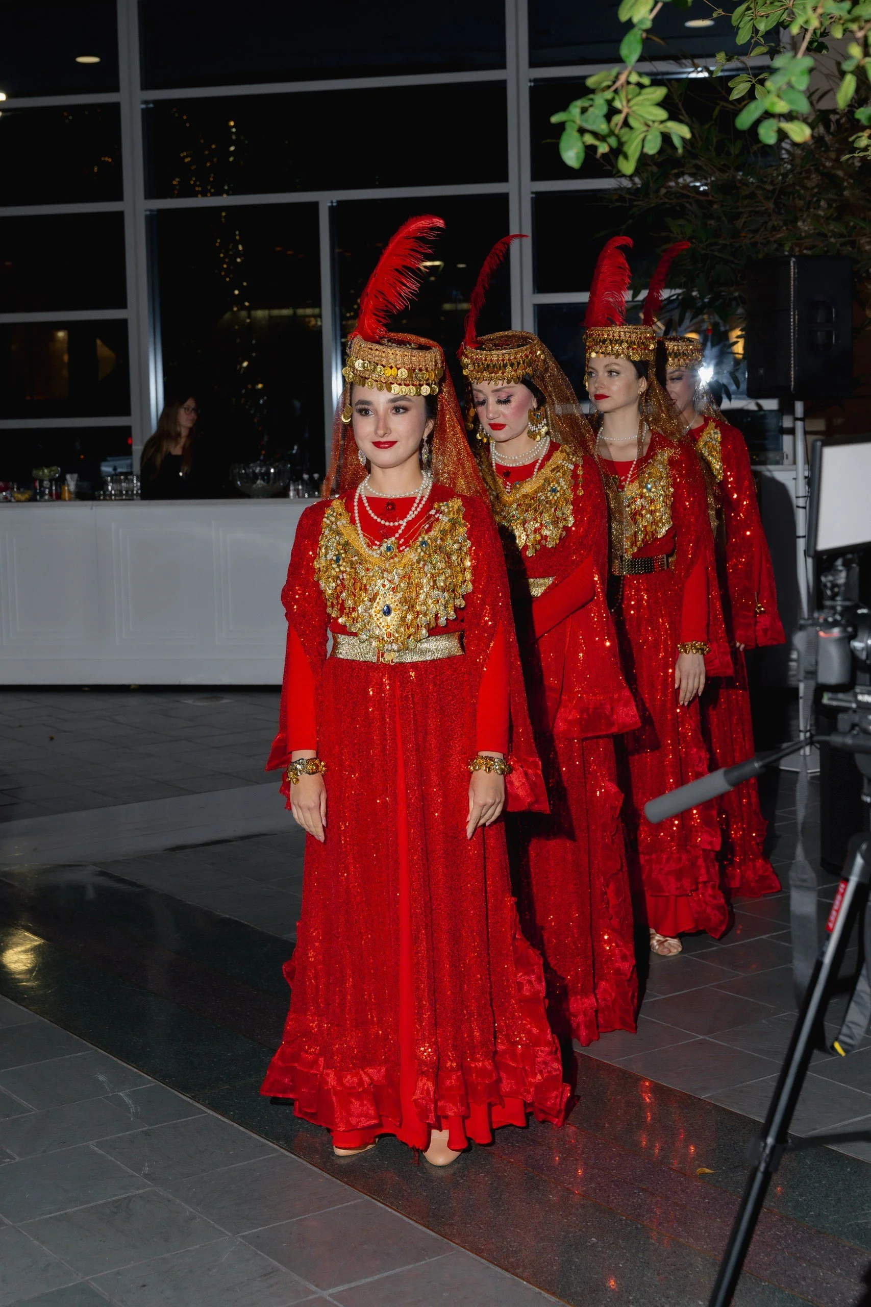 Group of women in traditional red evening gowns and gold jewelry, wearing tall red feathered headdresses, walking in a line at a formal event, with a building and indoor decor in the background.