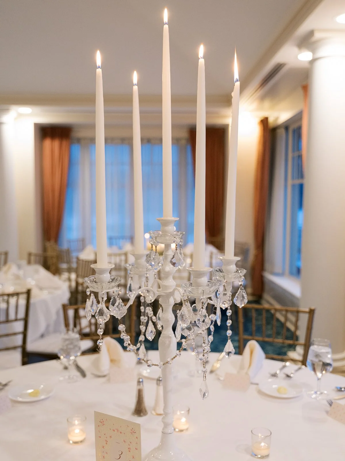 Elegant dining room with a crystal chandelier centerpiece with lit candles in a formal setting, decorated with wedding or event table arrangements.
