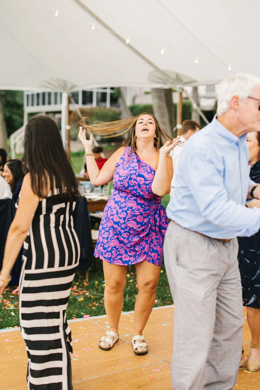 Woman in a pink and blue dress dancing at an outdoor event under a tent, with other people around.