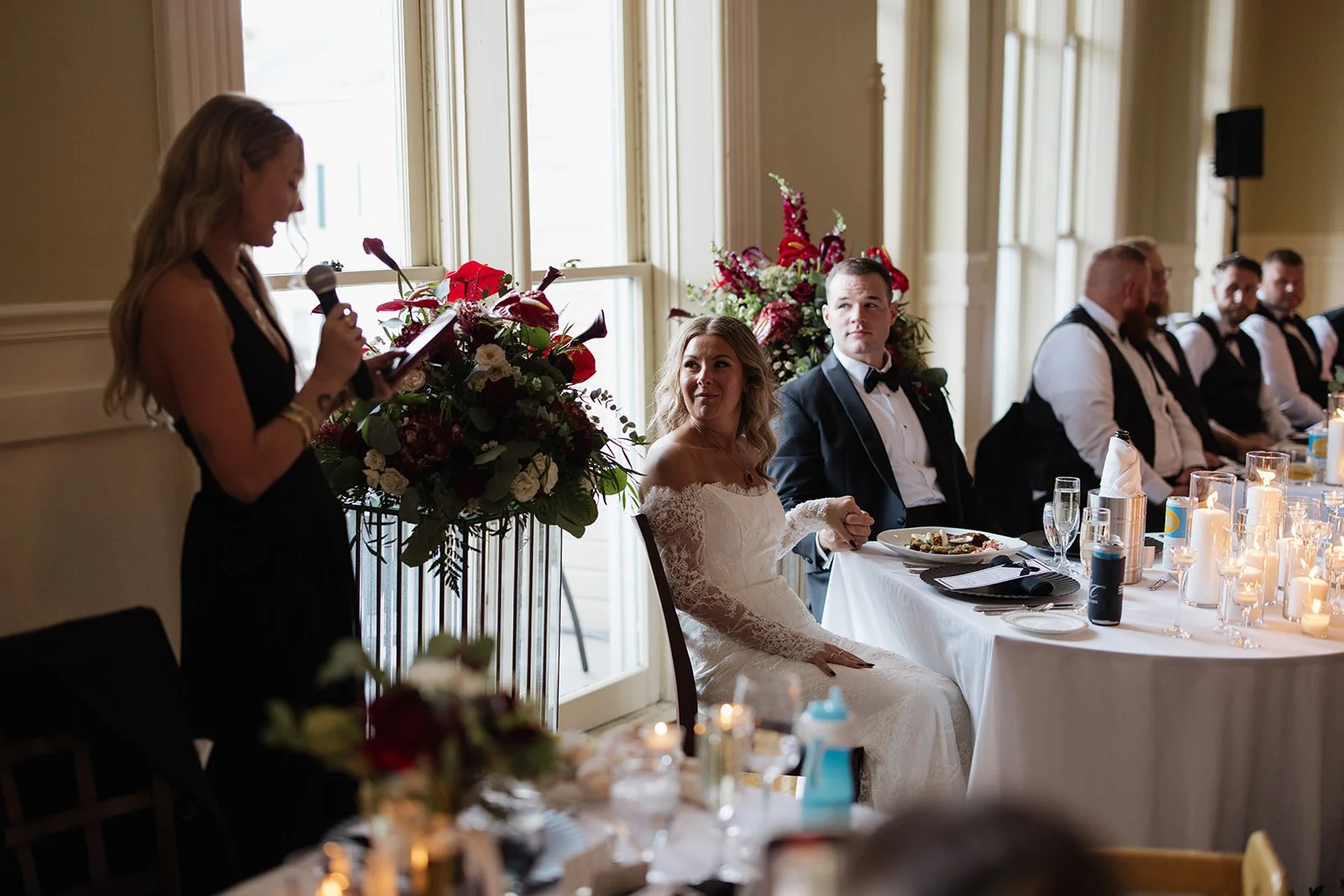 Bride in white wedding dress sitting at a reception table with wedding guests, listening to a woman giving a speech, with floral arrangements and candles on the table.
