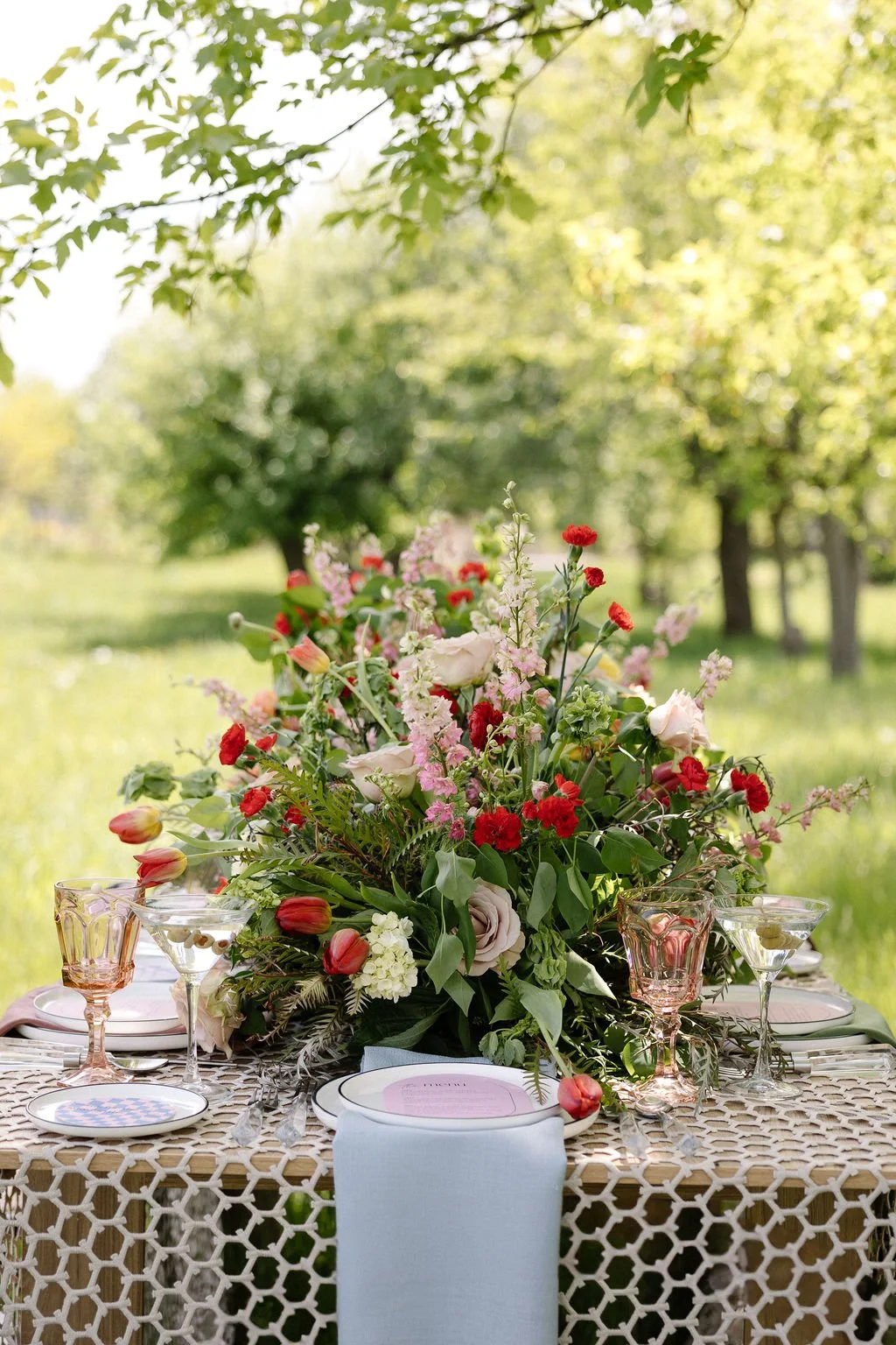 A table set for an outdoor event with a large floral centerpiece, pink and red flowers, glassware, and plates, in a green, tree-filled park setting.