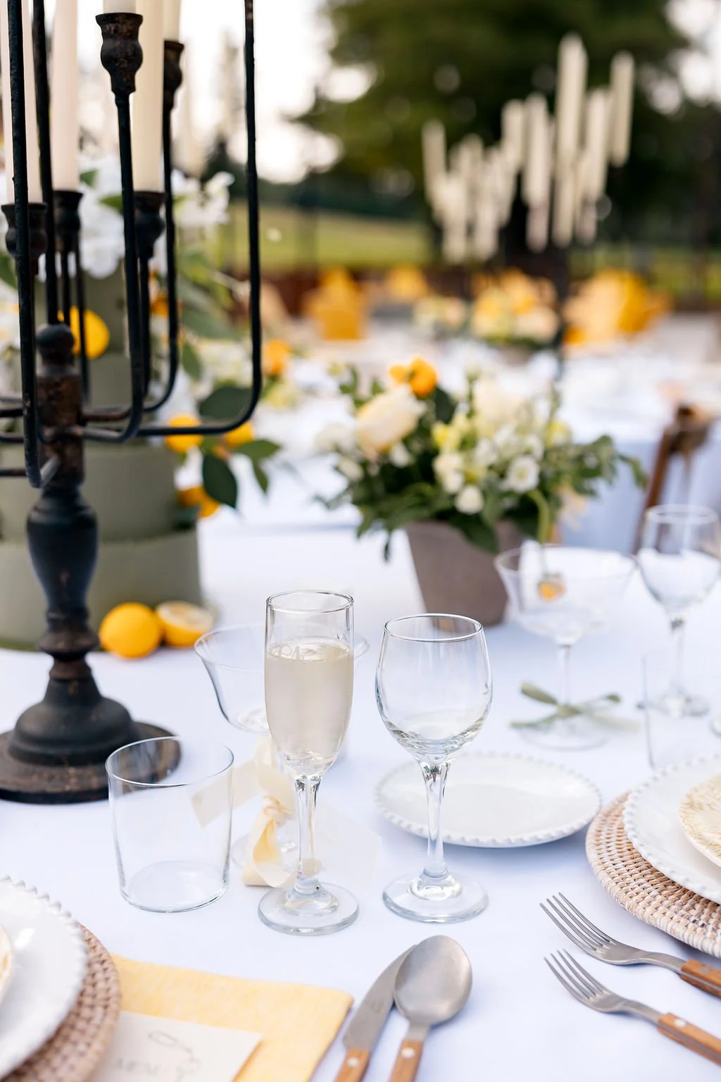 A decorated outdoor table with glassware, flowers, candles, and lemons for an event or celebration.