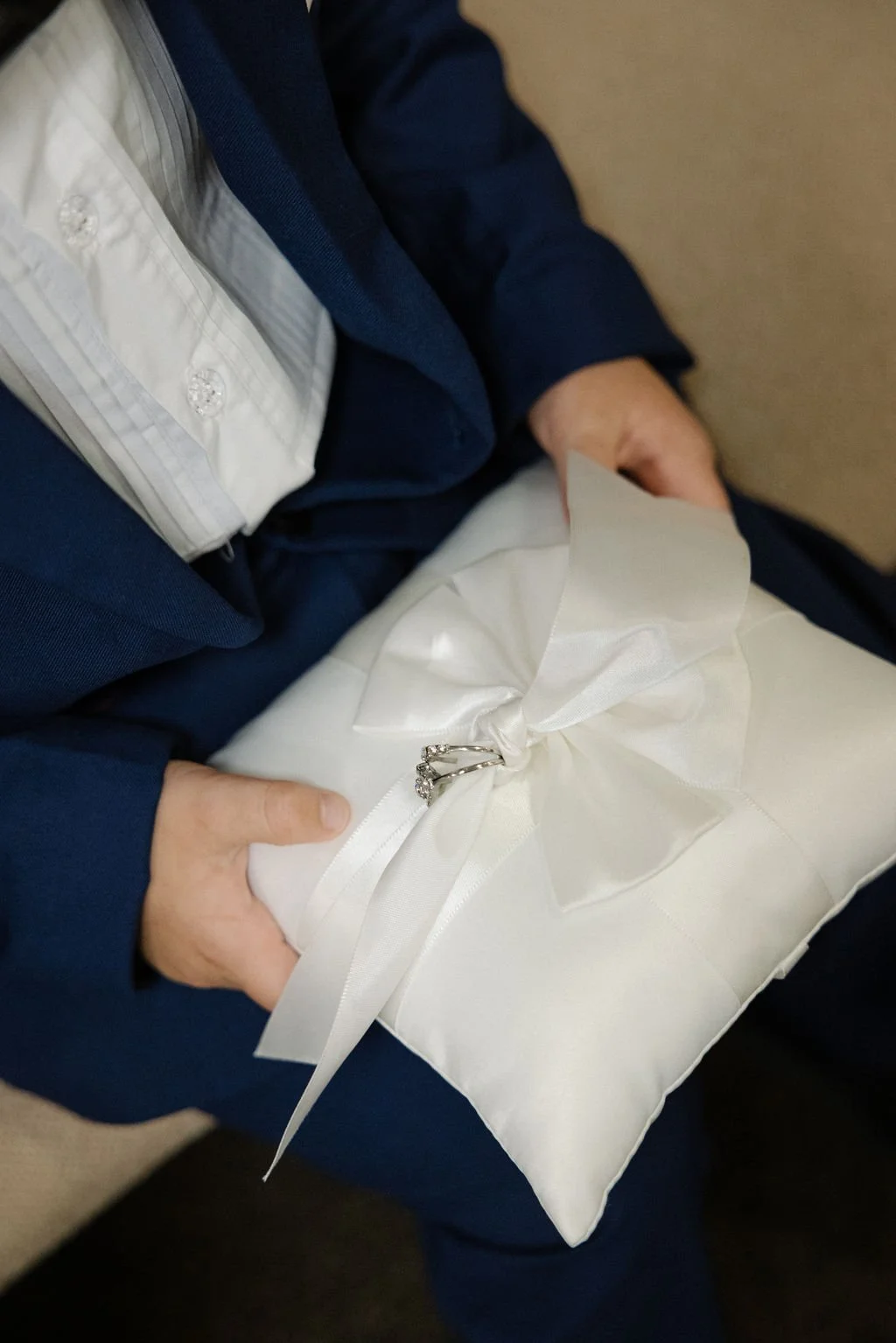 Person wearing a navy suit and white shirt holding a wrapped gift with a white satin ribbon and ring on top.