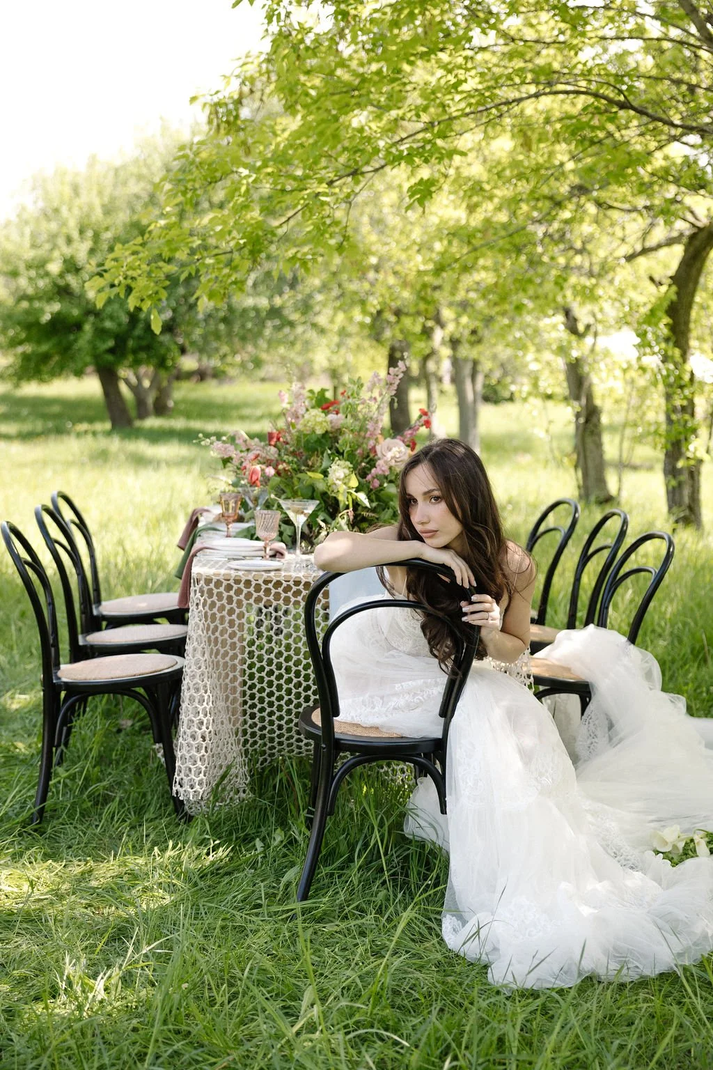 A woman in a wedding dress sitting on a black chair outdoors near a decorated table with floral arrangements and glasses, surrounded by green grass and trees.