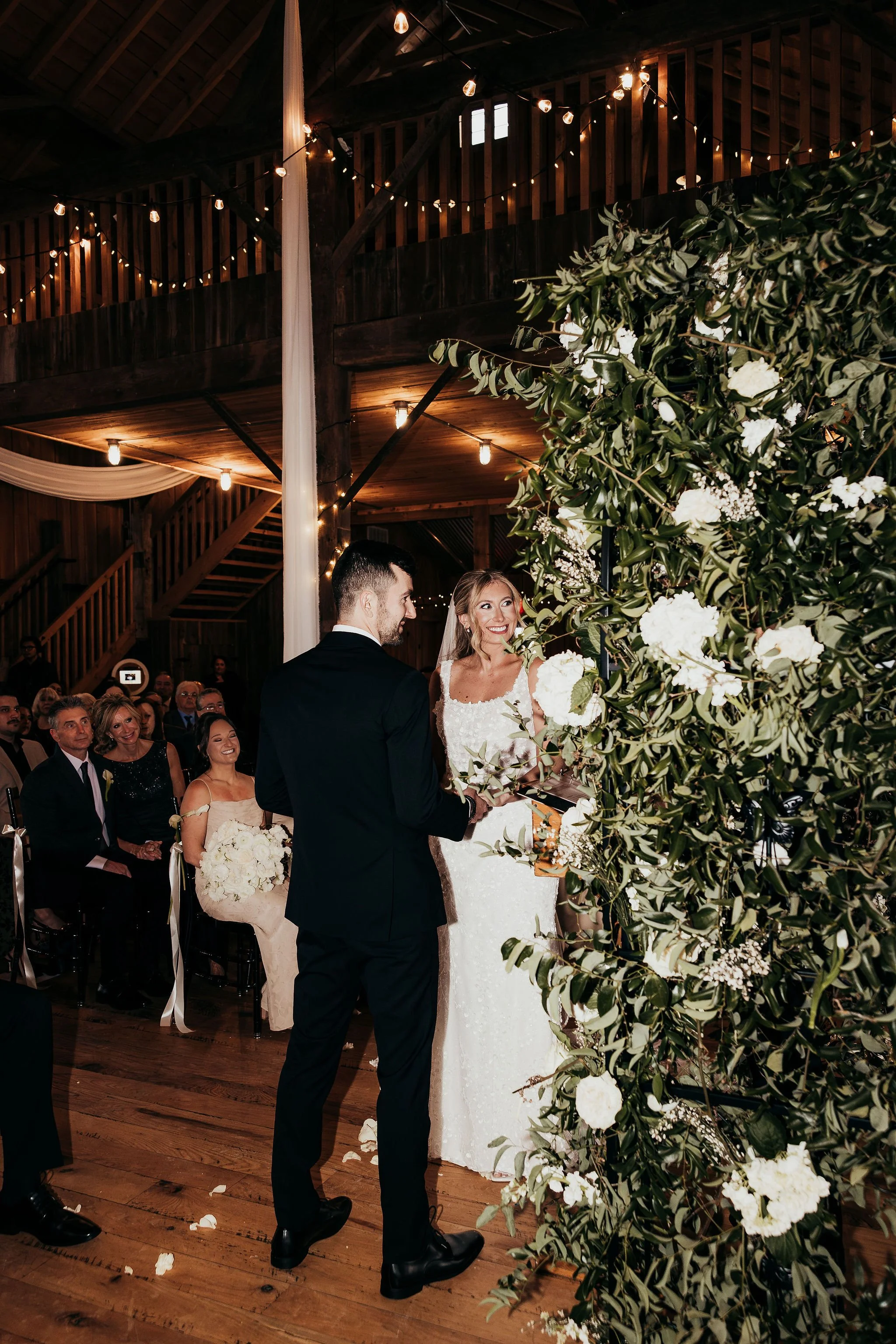 A wedding ceremony inside a rustic barn with string lights, featuring a bride in a white gown and a groom in a suit, standing in front of a large floral arrangement with white flowers and greenery.