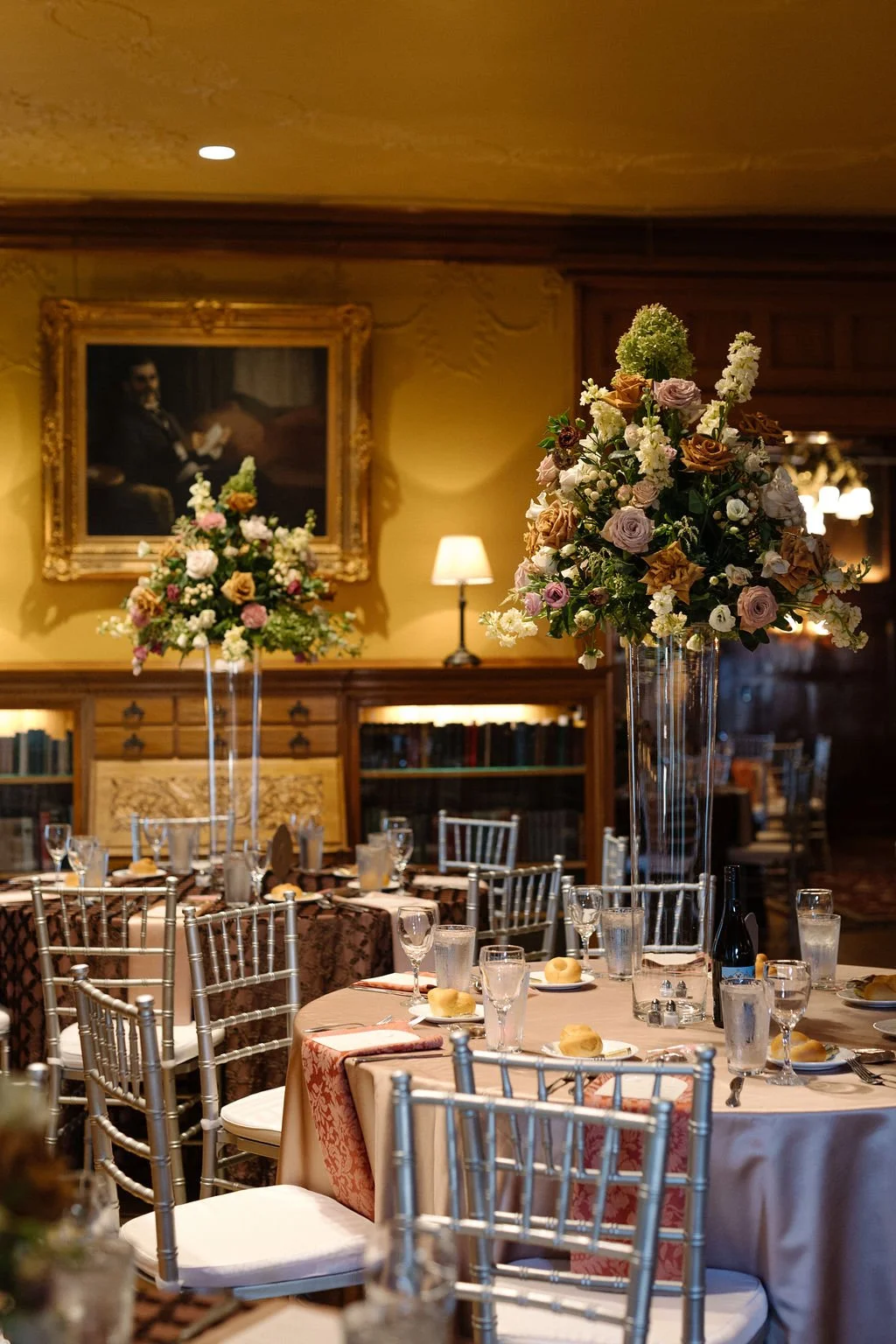 Elegant dining room decorated for a special event with tall floral centerpieces of roses and greenery on the tables, silver chairs, and a portrait of a man reading a book hanging on the wall.