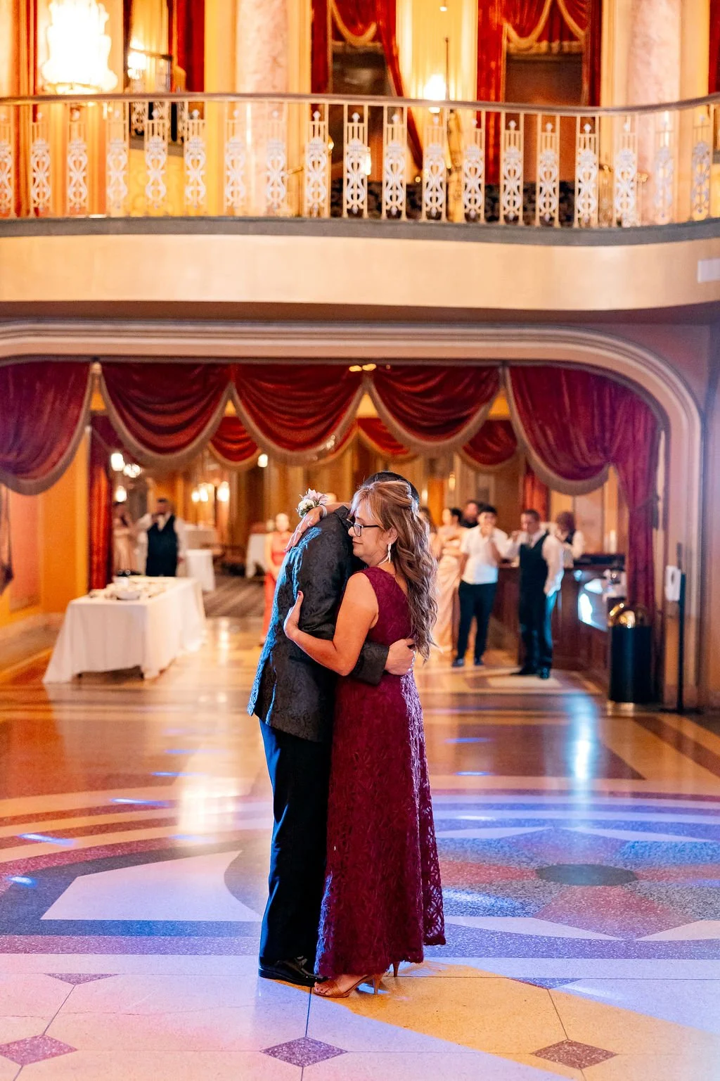 Couple hugging on the dance floor at a wedding reception with red curtains and other guests in the background.