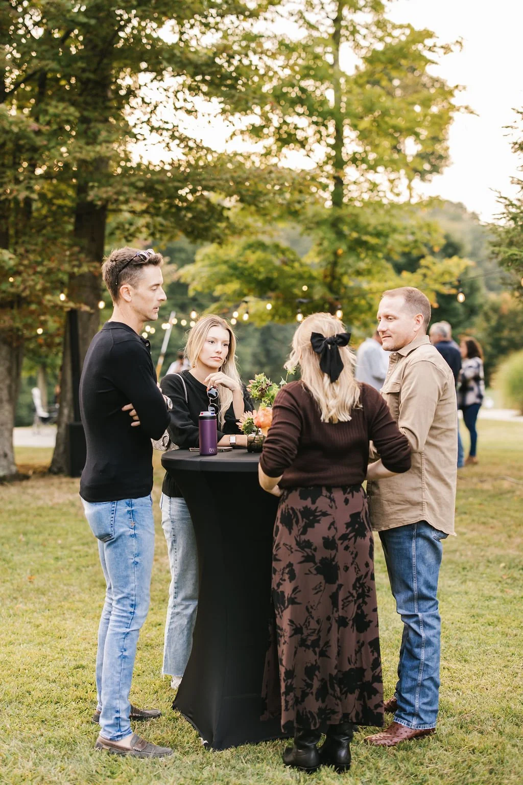 Four people gathered around a standing table outdoors during early evening at a social event, with trees and string lights in the background.