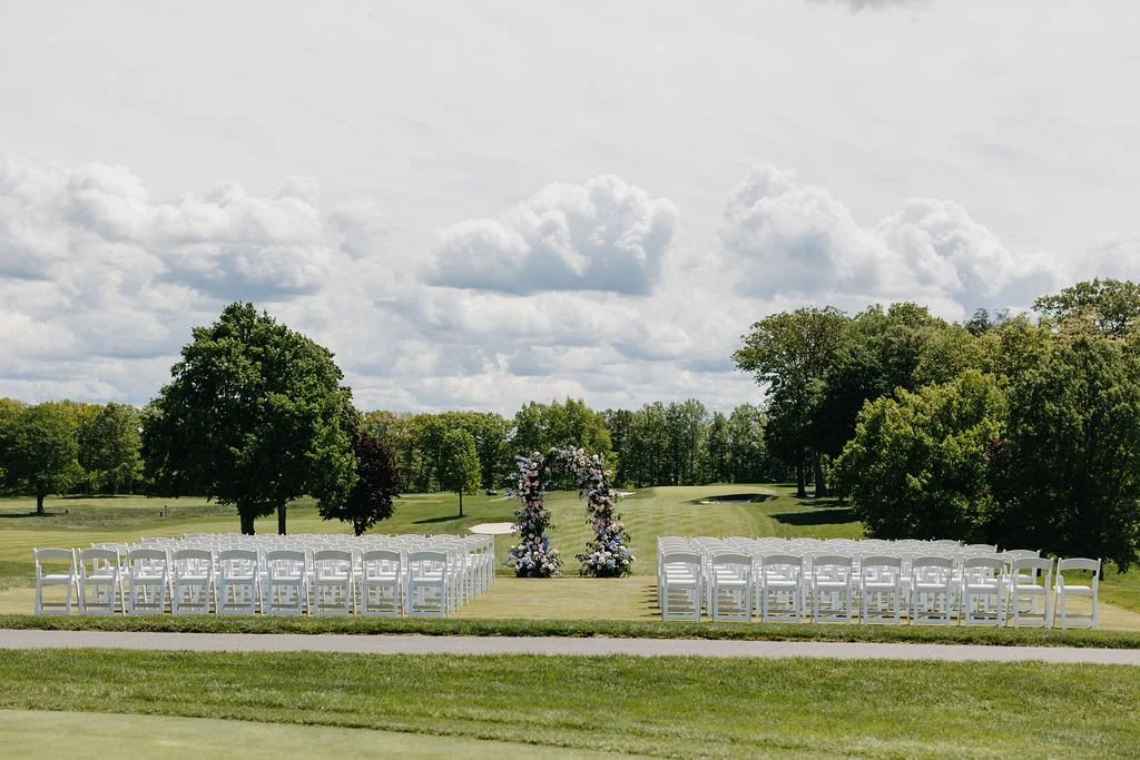 An outdoor wedding ceremony setup on a grassy field with rows of white chairs facing a floral arch, surrounded by trees and under a cloudy sky.