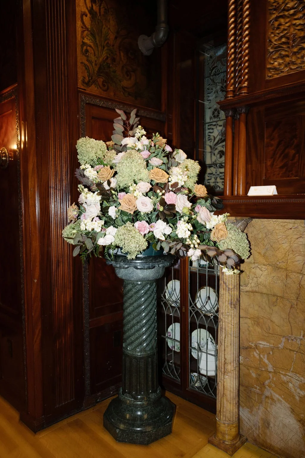 A large floral arrangement with white, pink, and beige flowers, including roses and hydrangeas, in a tall black pedestal vase inside a room with wood-paneled walls and a marble fireplace.