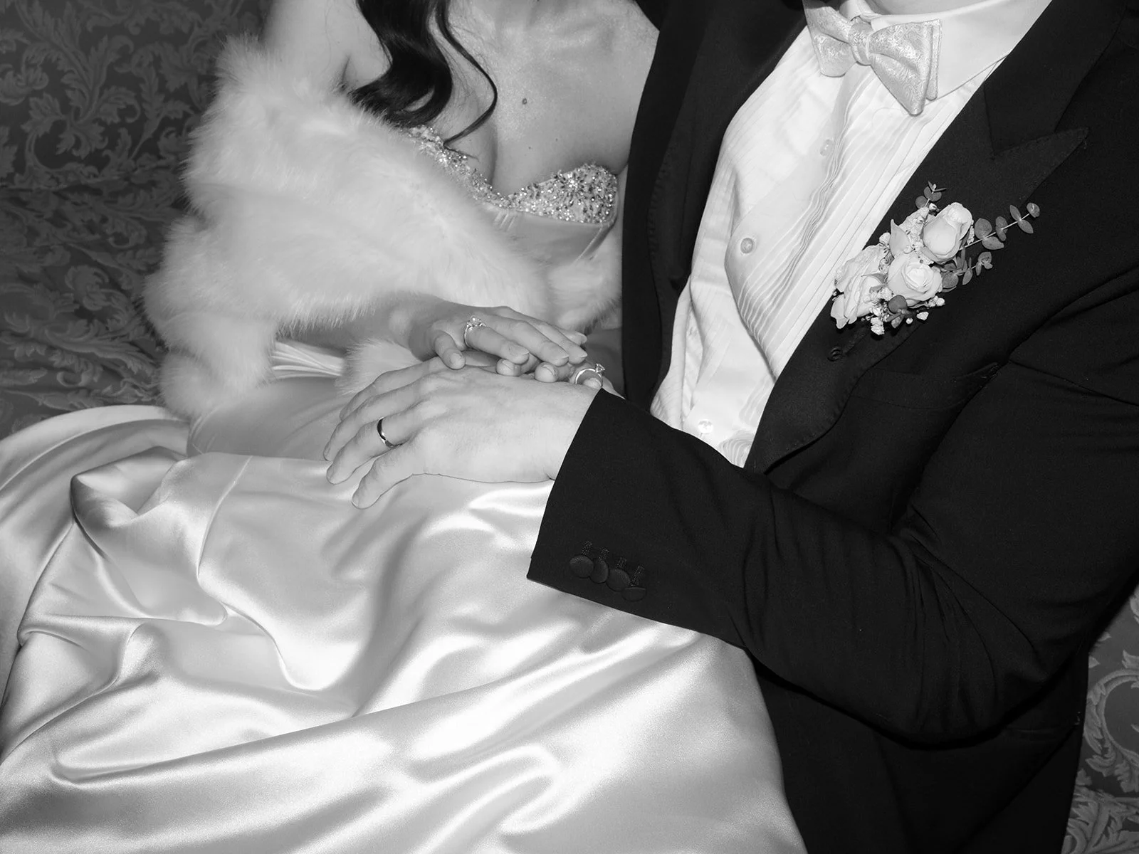 A close-up black and white photo of a bride and groom holding hands during their wedding. The bride is wearing a dress with a beaded neckline and a fur stole, with rings on her fingers. The groom is in a tuxedo with a bow tie and boutonniere, holding