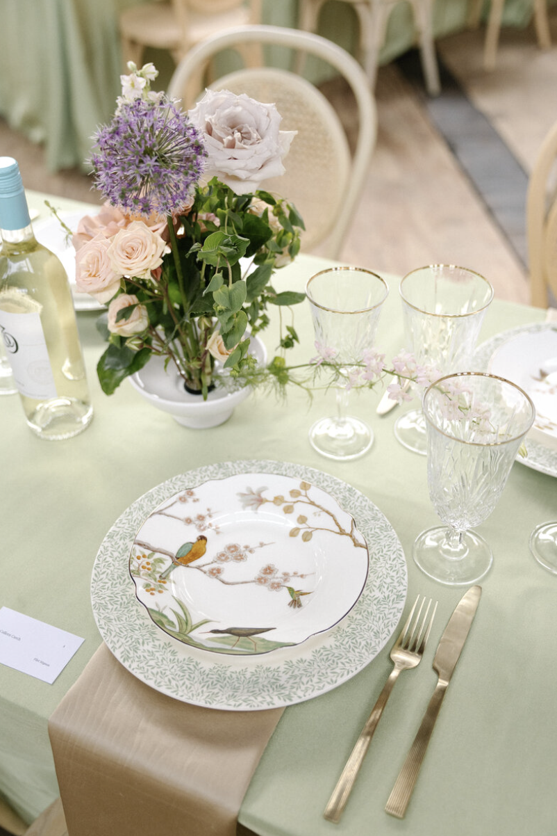 A table setting with a floral centerpiece, decorative plates featuring bird and flower designs, gold-trimmed glasses, gold cutlery, a light green tablecloth, and a beige table runner.