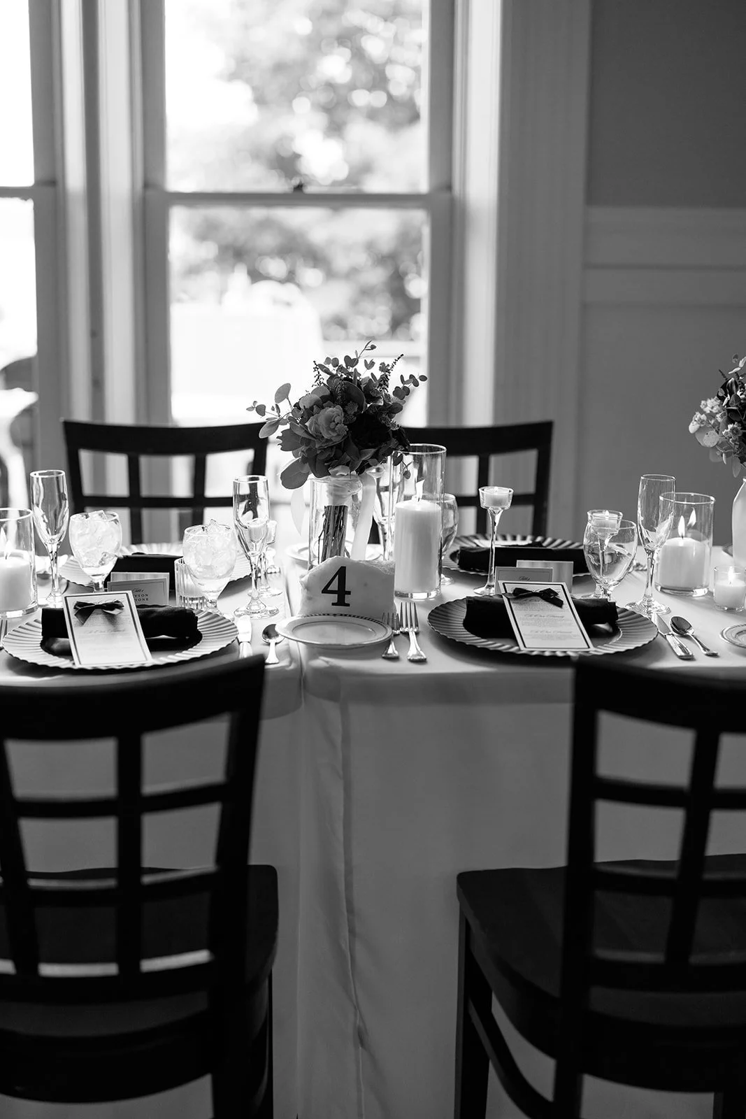 A table set for an event with floral centerpiece, candles, wine glasses, black napkins, and printed menus in a room with large windows.