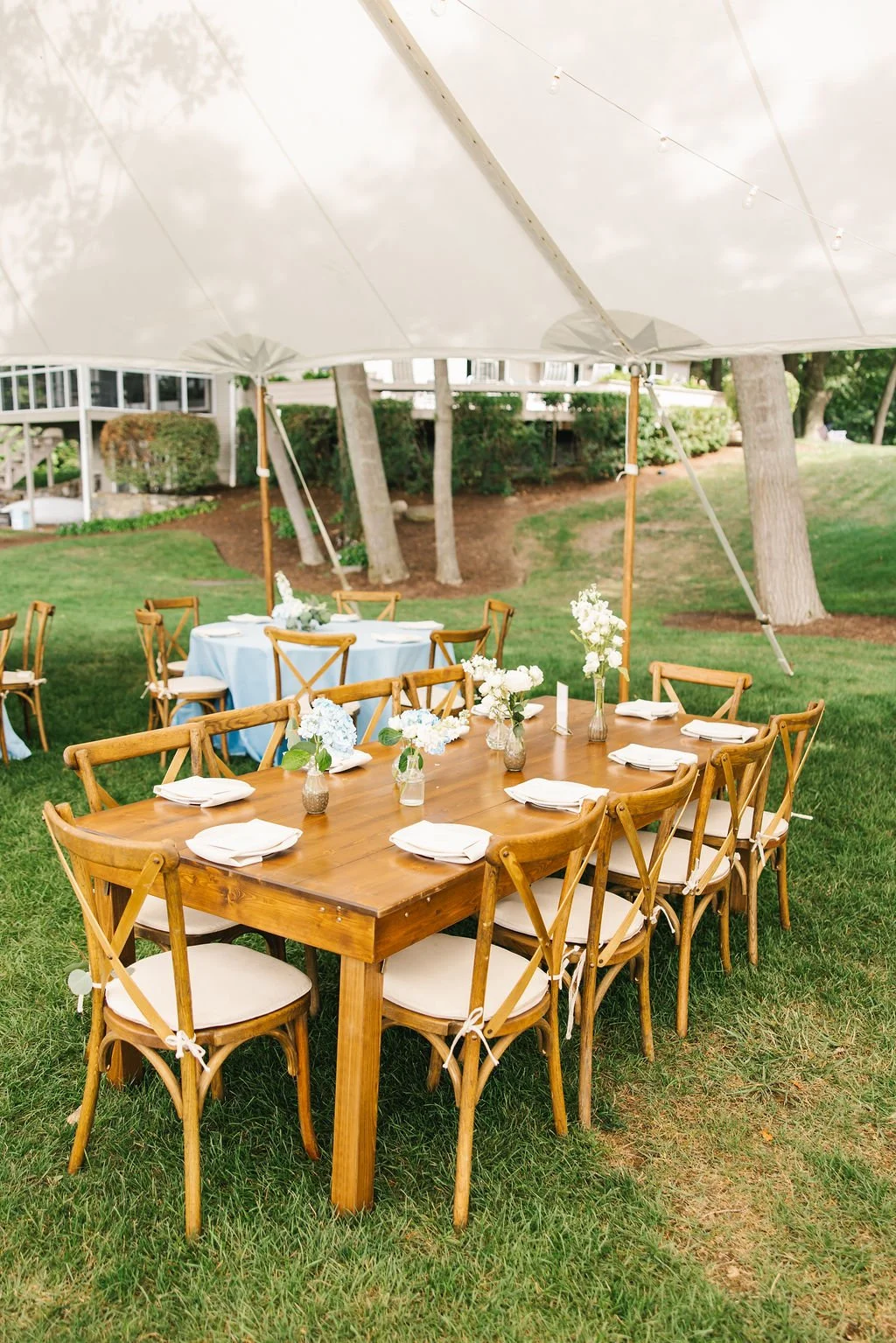 Outdoor wedding or event seating with a large wooden table set with white napkins and vases of white flowers, under a beige canopy on green grass, with additional tables and trees in the background.