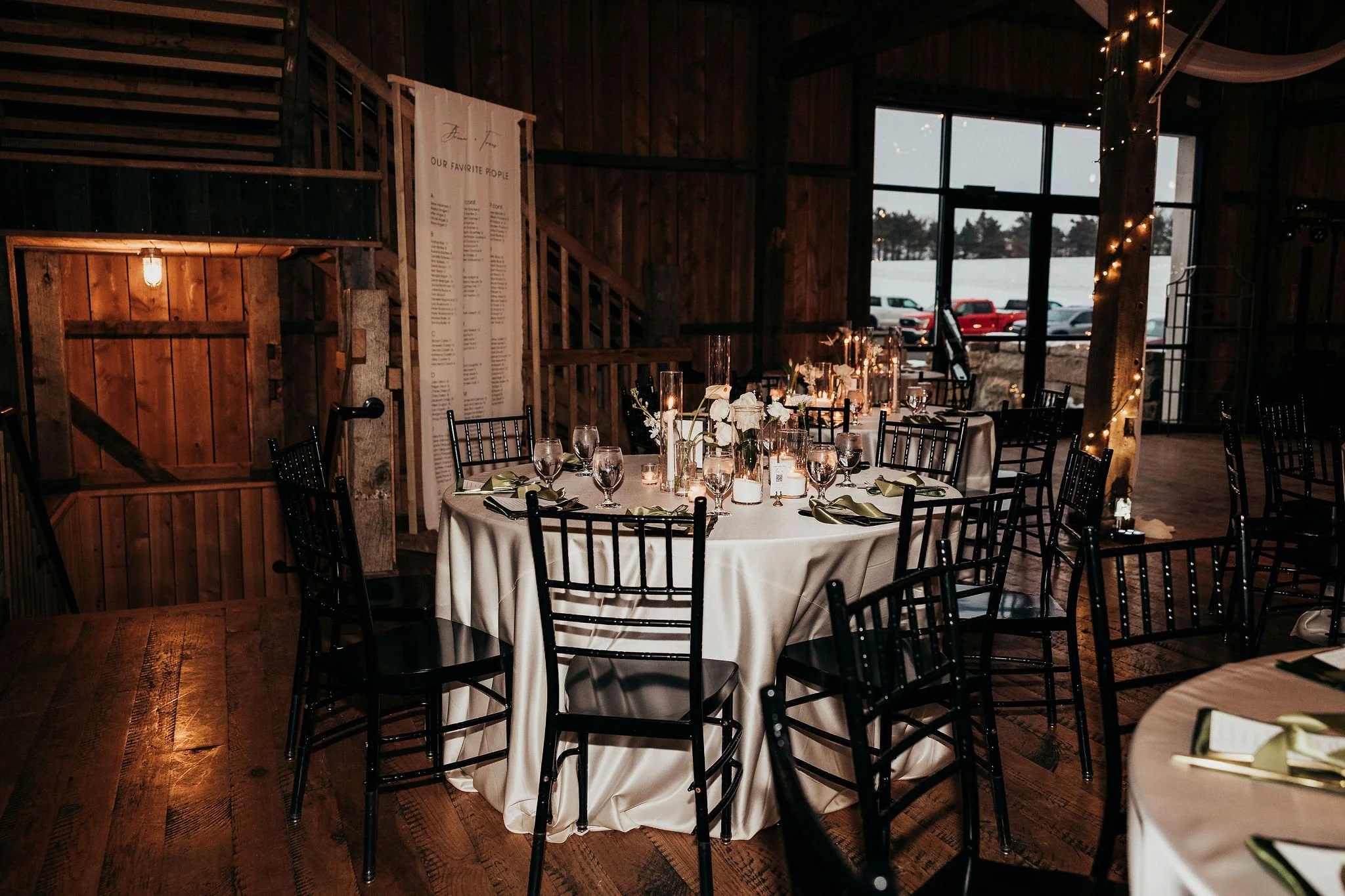 Indoor rustic wedding reception with round table set with glassware and flowers, surrounded by black chairs, large window showing a snowy landscape, wooden beams, and string lights.