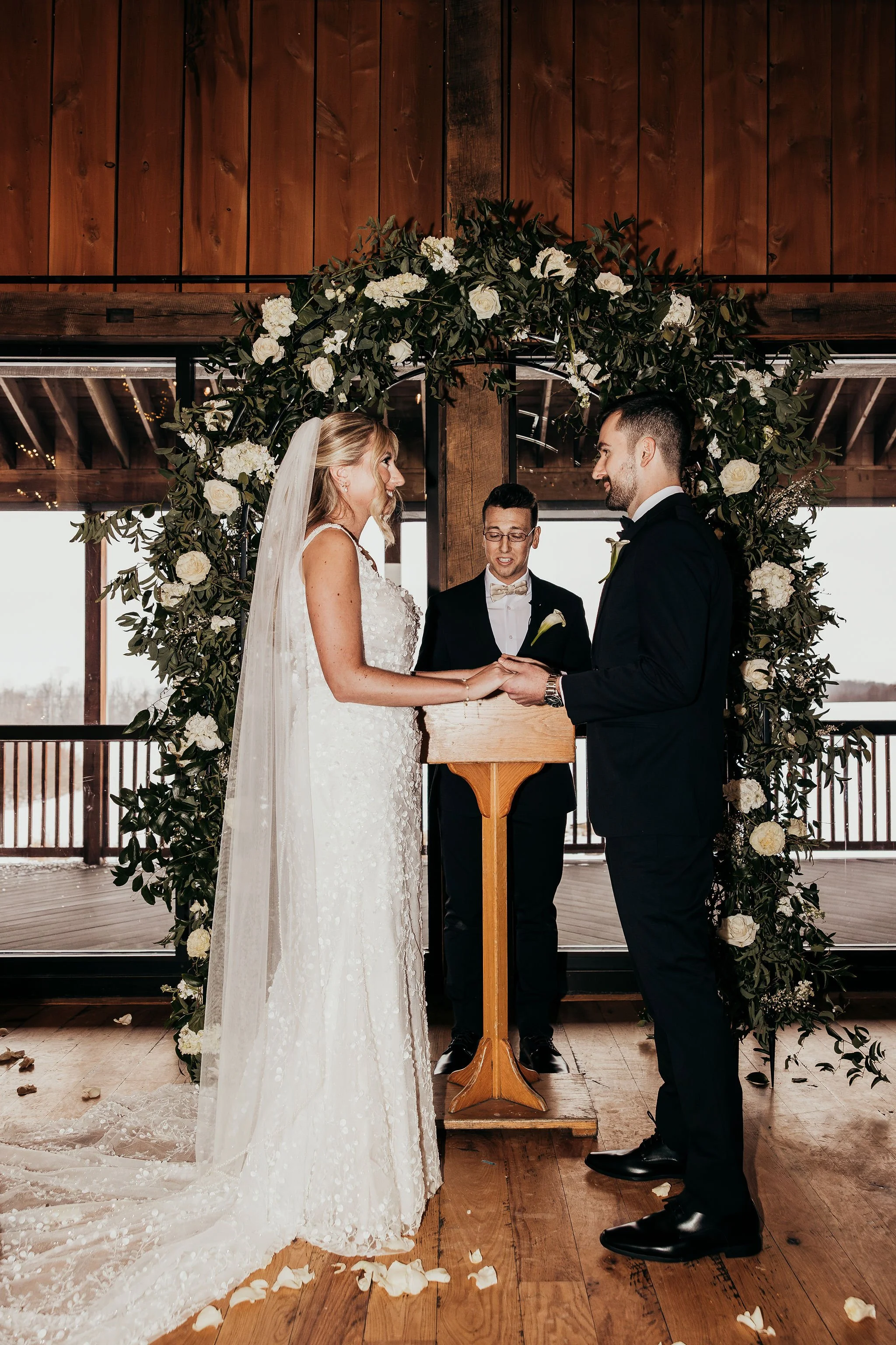 A bride and groom exchanging vows during a wedding ceremony, standing under a floral arch with an officiant.