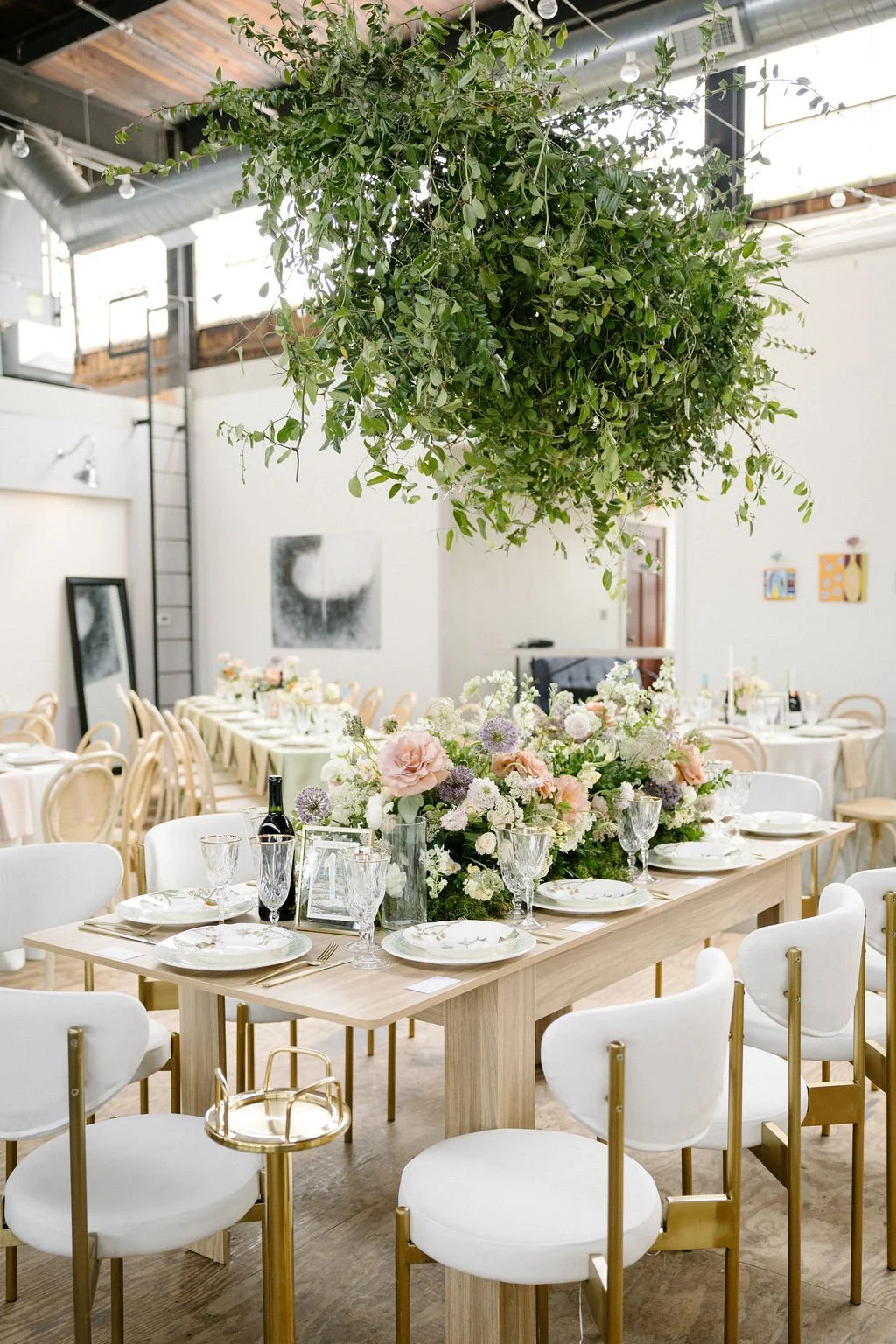 Elegant dining setup with a hanging greenery arrangement and a floral centerpiece on a wooden table surrounded by white chairs with gold accents.