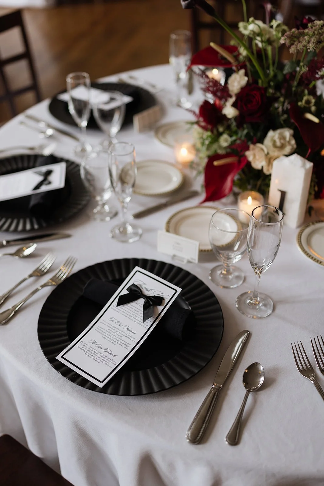 A formal dining table set with black and white dinnerware and glassware, a floral centerpiece with red and white flowers, candles, and place cards, for an elegant event or wedding.