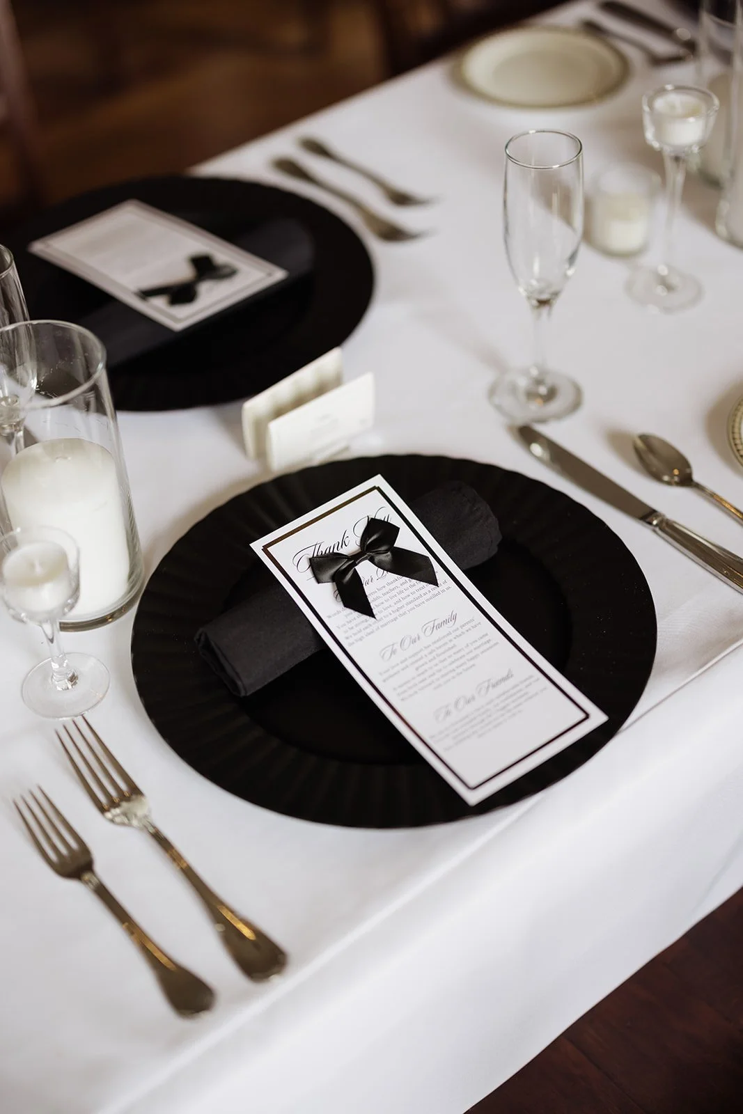 Elegant wedding table setting with black and white color theme, includes black dinner plate, black napkin with a thank you card tied with a black ribbon, champagne flute, candles, silverware, and a white tablecloth.