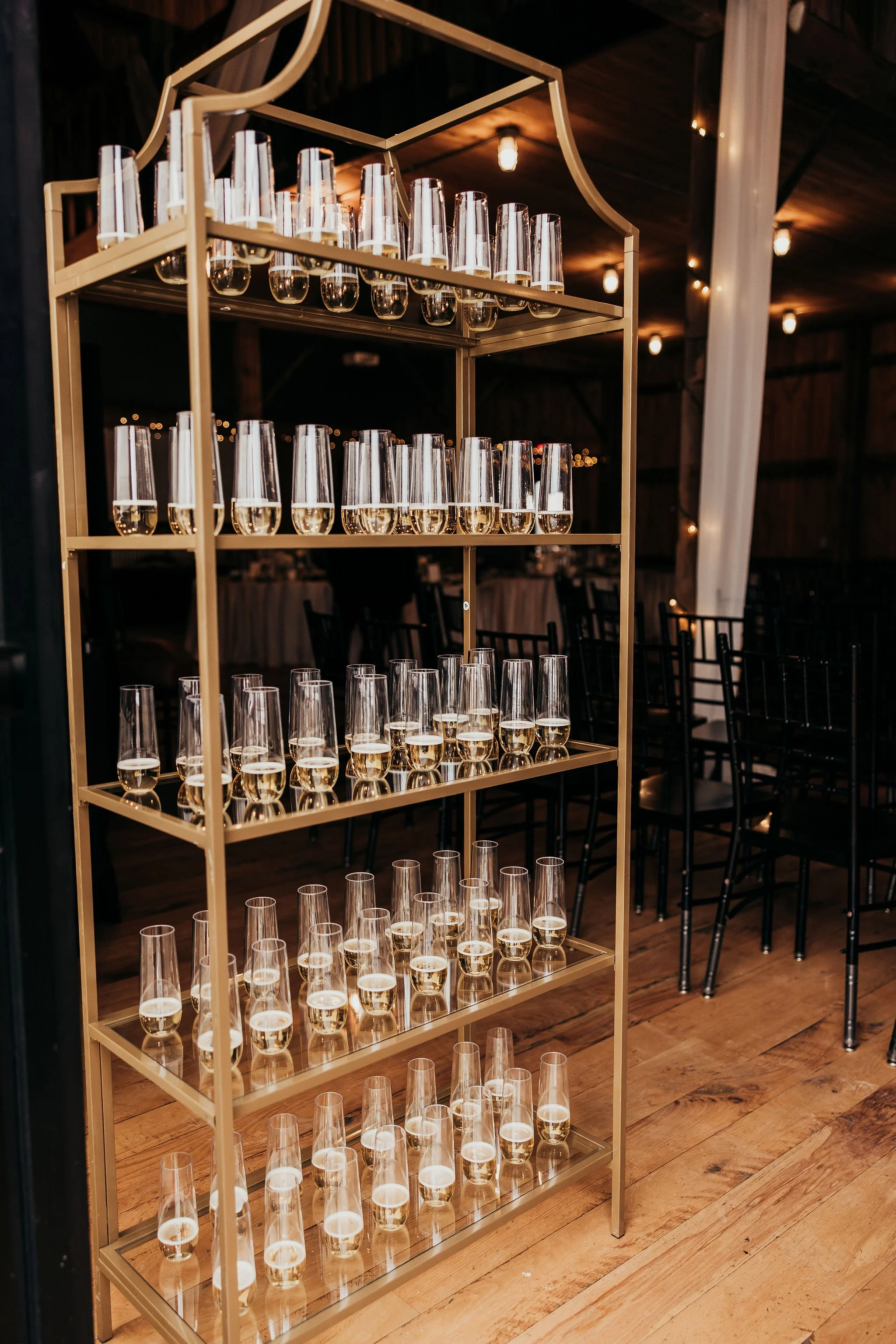A gold-colored metal shelving unit filled with champagne glasses, most of which are half full, in a dimly lit venue with wooden floors and chairs in the background.