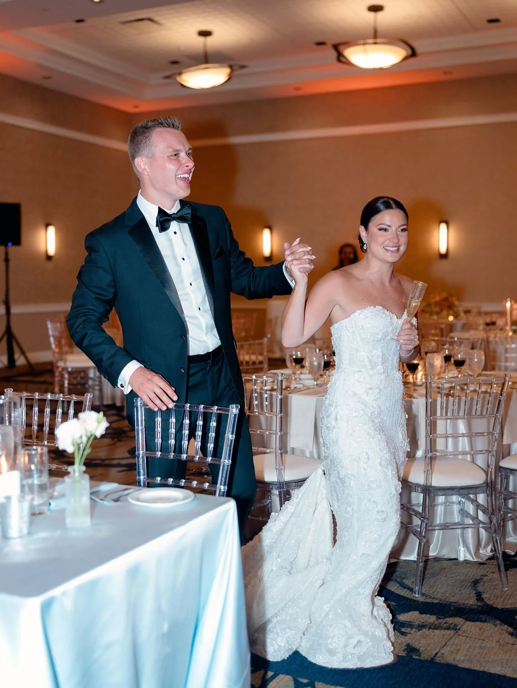 A bride and groom dancing at their wedding reception. The groom is wearing a tuxedo and the bride a white wedding dress, holding hands and smiling.