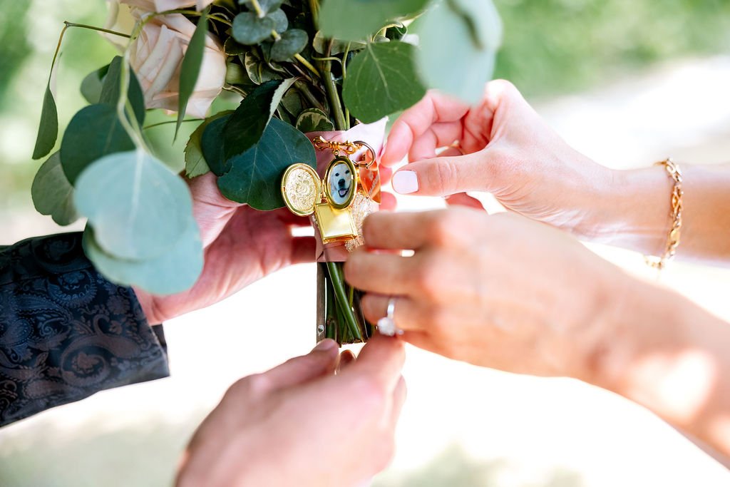 Two hands attaching a small bouquet of green leaves and white roses, decorated with a gold pin and a portrait charm, at an outdoor event.
