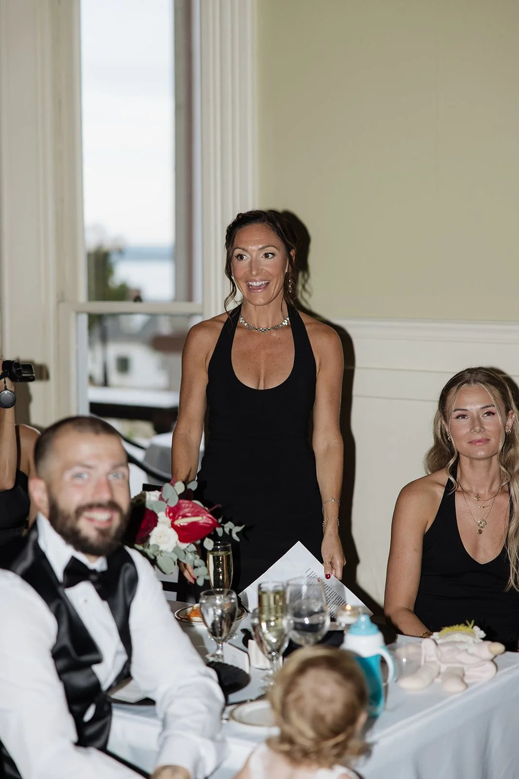A woman in a black dress standing at a banquet table, smiling, with guests in formal attire seated around.