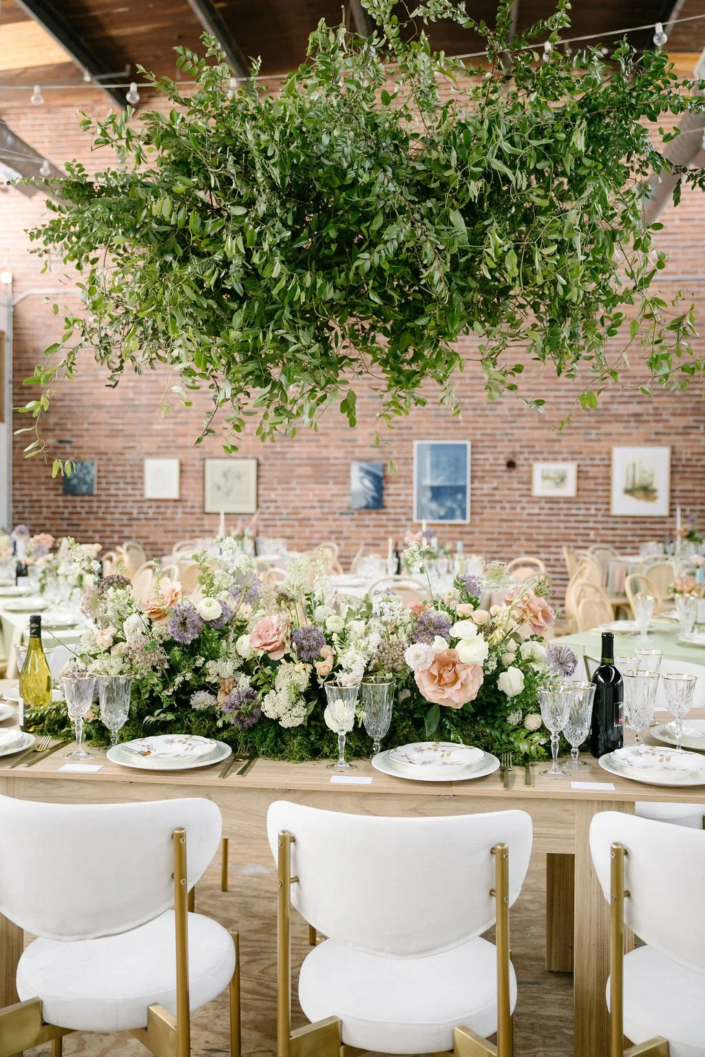 Elegant event setup with a long wooden table decorated with a lush floral centerpiece, surrounded by white chairs, in a venue with a brick wall and framed artwork, and a large hanging green plant above.