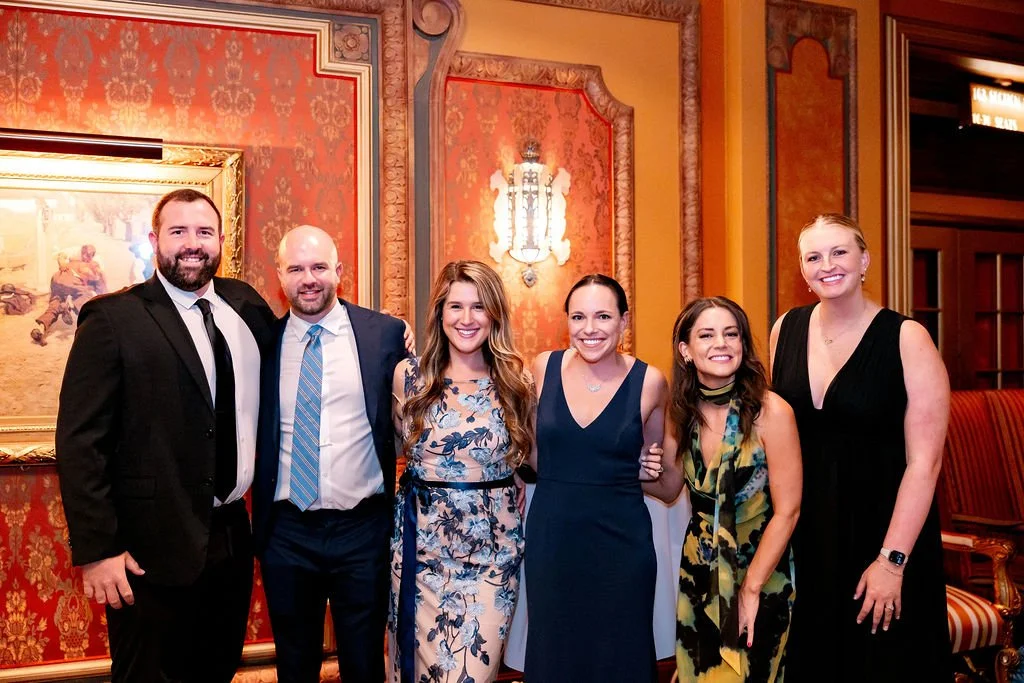 Group of six people dressed in formal attire smiling in an elegant, ornately decorated room.