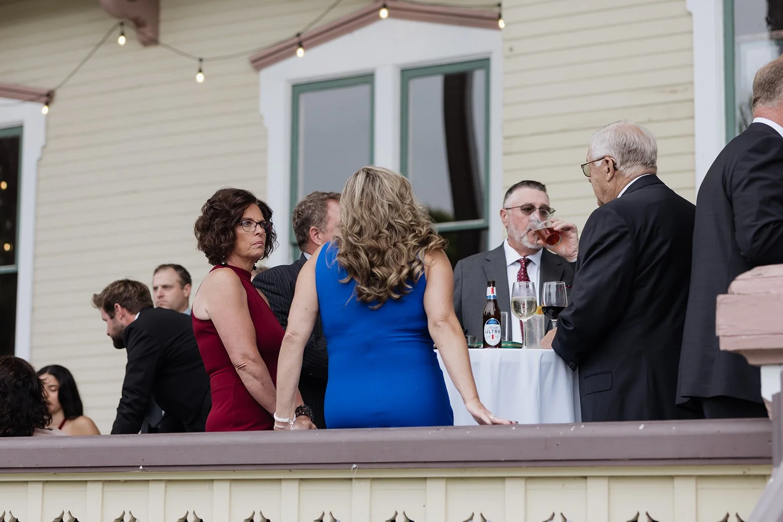 People standing and socializing at an outdoor gathering or party on a balcony, with drinks and conversing.