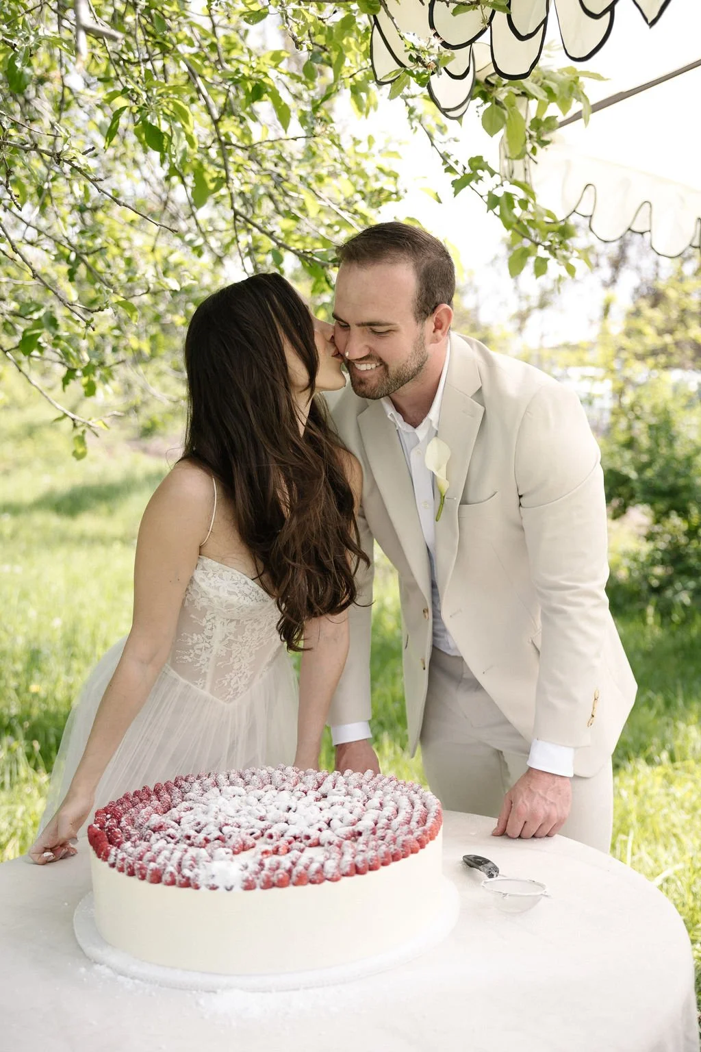 A newlywed couple kissing outdoors at their wedding reception, with a large strawberry cake on the table in front of them, under a canopy of leafy trees.