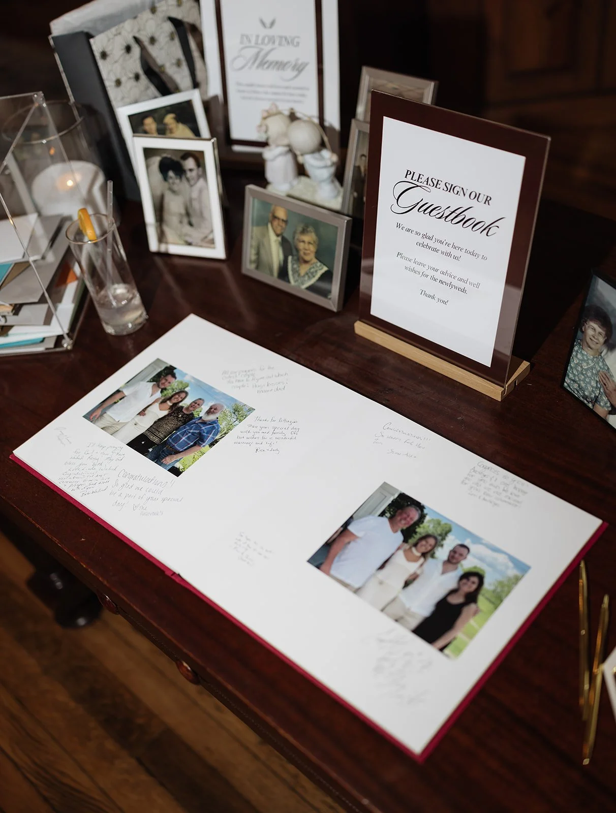 A guestbook display at a wedding reception with framed photos of the couple and family, a sign inviting guests to sign the guestbook, and an open guestbook with photos and handwritten messages.