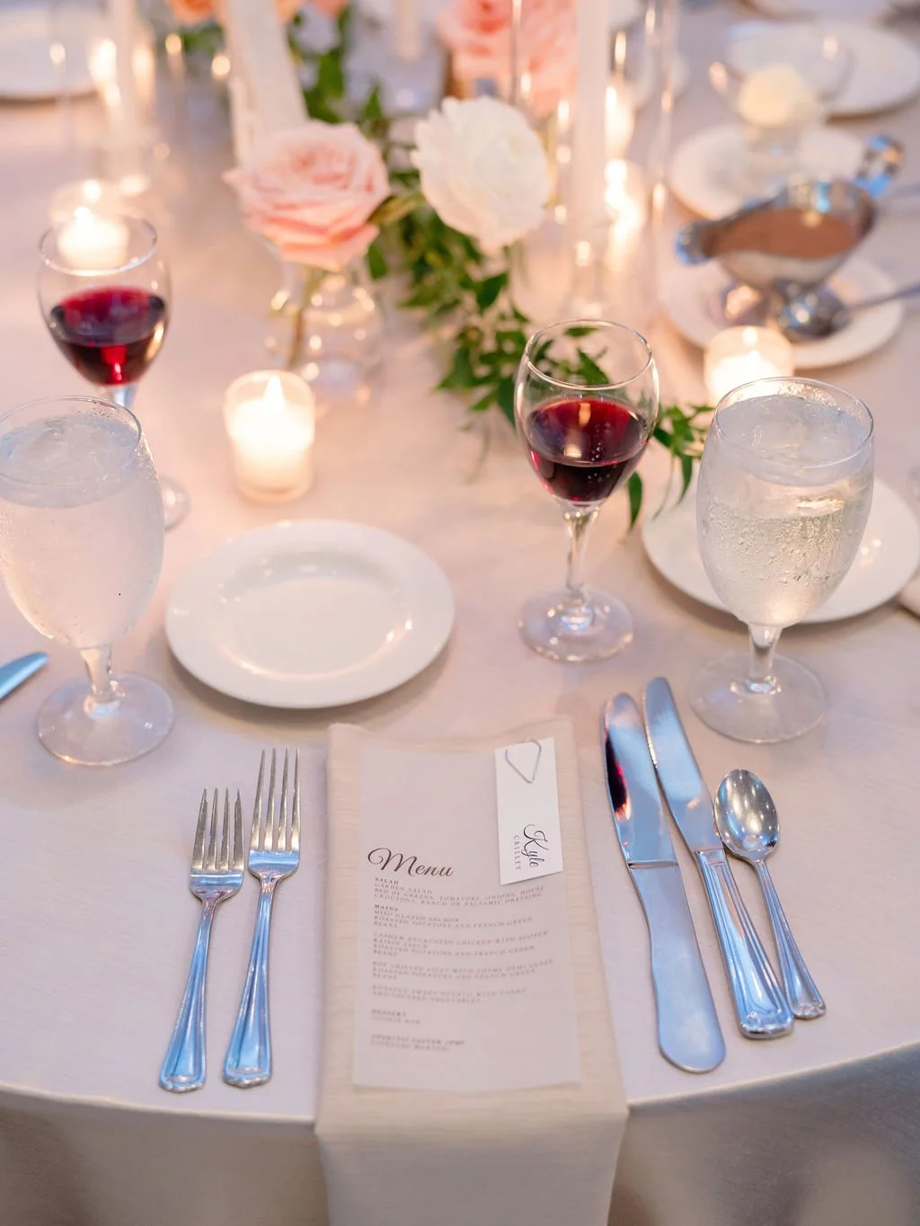 Elegant table setting with wine glasses, water glasses, cutlery, a menu, candles, and floral centerpieces for a formal event.