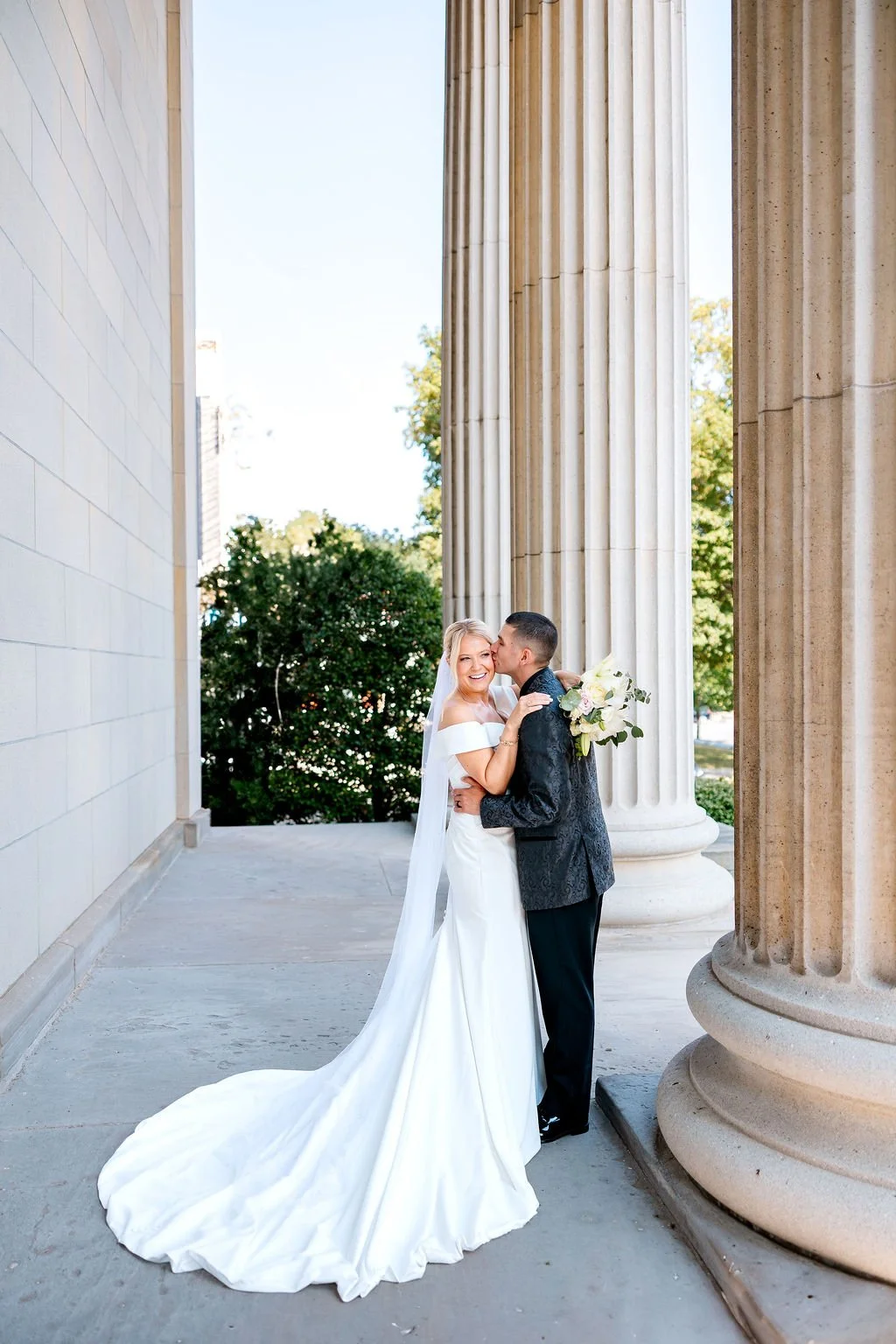 A bride and groom sharing a kiss outside a building with tall classical columns. The bride wears a white off-the-shoulder gown with a long train, and the groom wears a dark patterned suit. The bride holds a bouquet of white flowers with greenery.