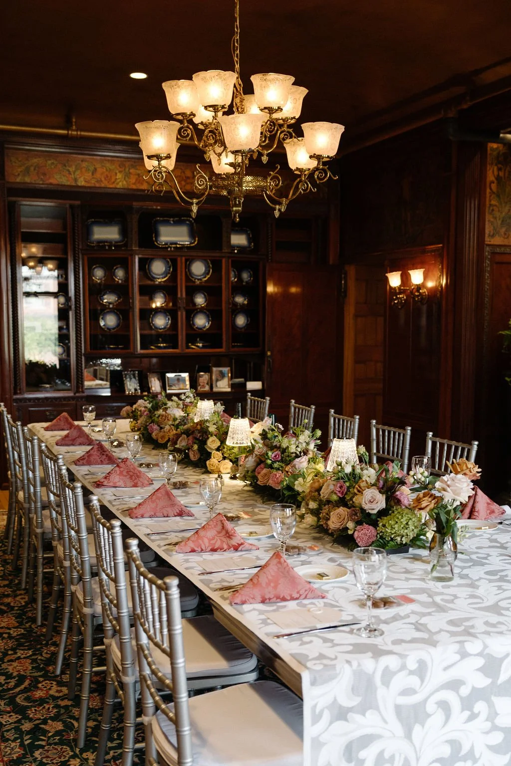 Elegant dining room with a long table set for a formal meal, decorated with a floral centerpiece, pink napkins, and glassware, under a chandelier with multiple light fixtures.