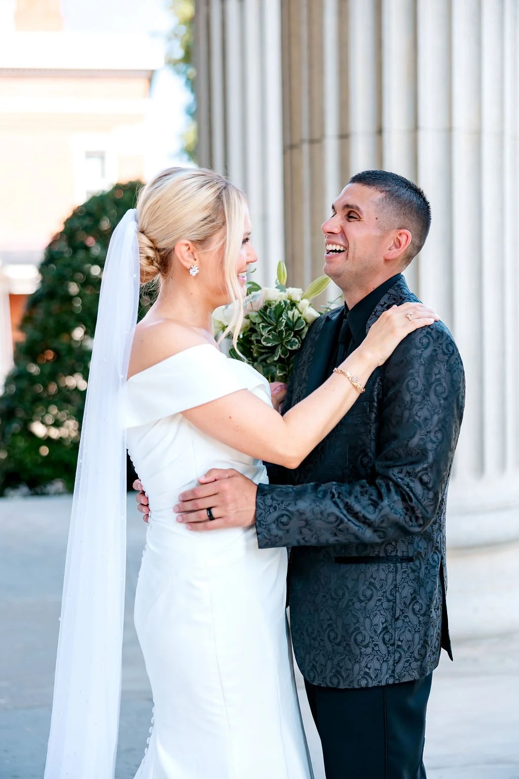 A bride and groom smiling and embracing outside a building with large columns, wedding day