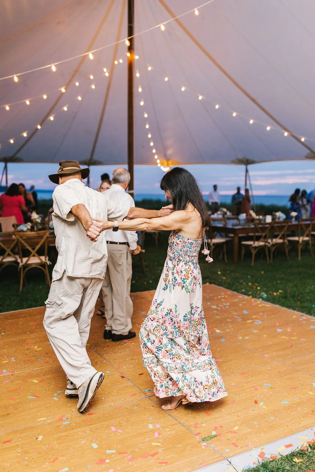People dancing on a wooden dance floor under a large tent decorated with string lights, with outdoors in the background, during a celebration or wedding reception.