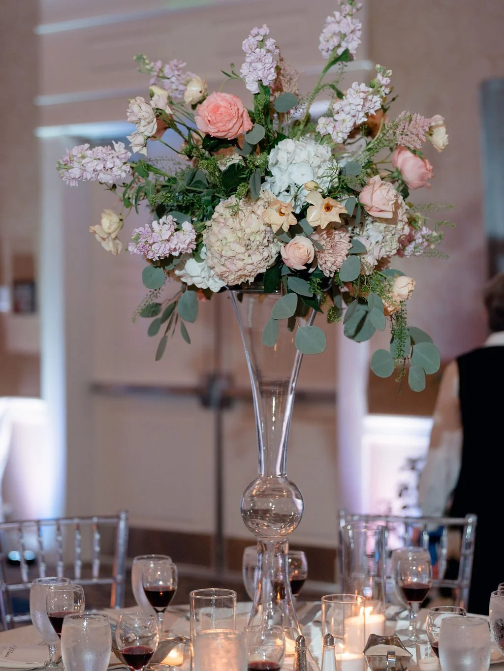 A tall glass vase with a large bouquet of pink and white flowers, green leaves, and eucalyptus, placed on a table set for a formal event with wine glasses, candles, and silverware.