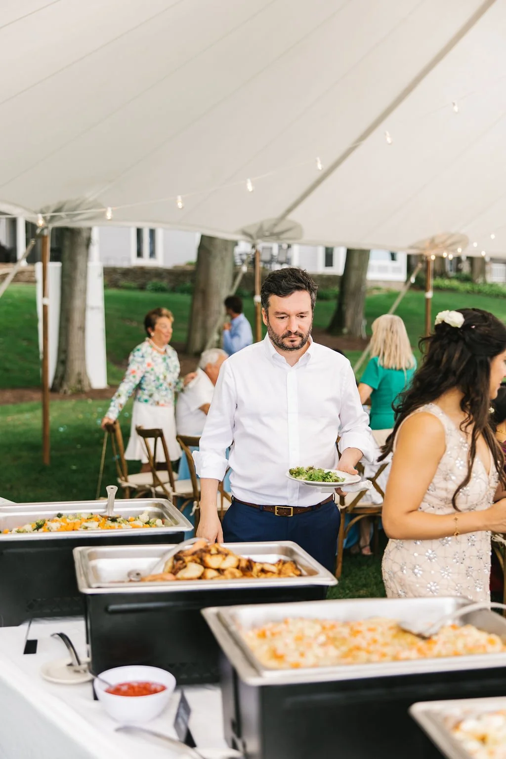 Man serving himself at an outdoor buffet table with dishes including salad, roasted chicken, and pasta at a gathering under a large white canopy with guests in the background.