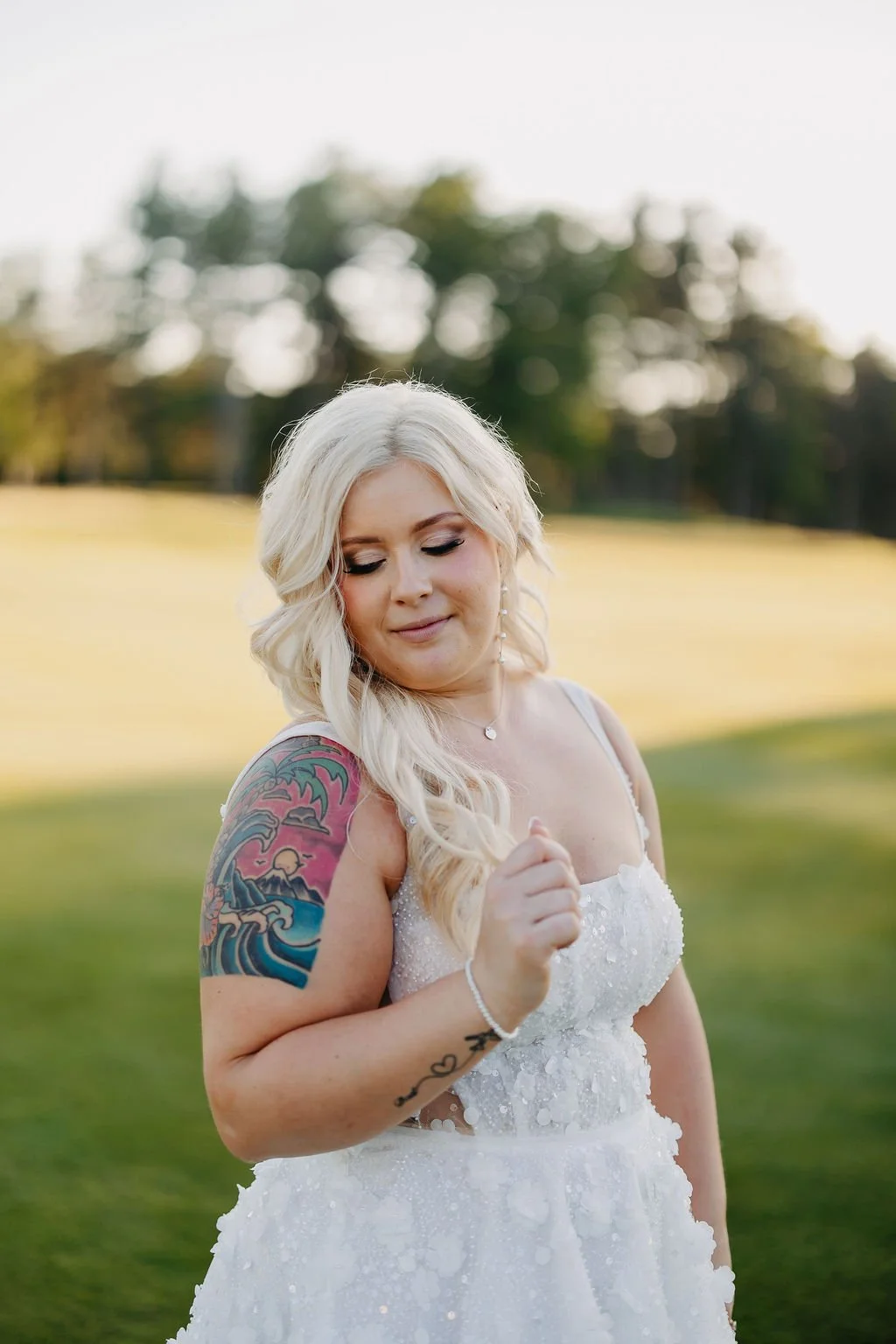 A woman with platinum blonde hair in loose waves, wearing a white dress, standing outdoors in a grassy area with trees in the background. She has a colorful tattoo sleeve on her left arm and is looking down with a gentle smile.