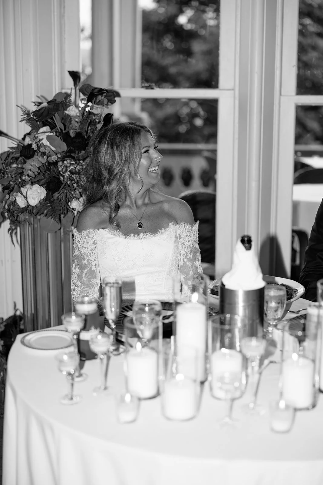 A woman in an off-the-shoulder lace wedding dress smiling at a formal event, sitting at a table with decorations and candles.