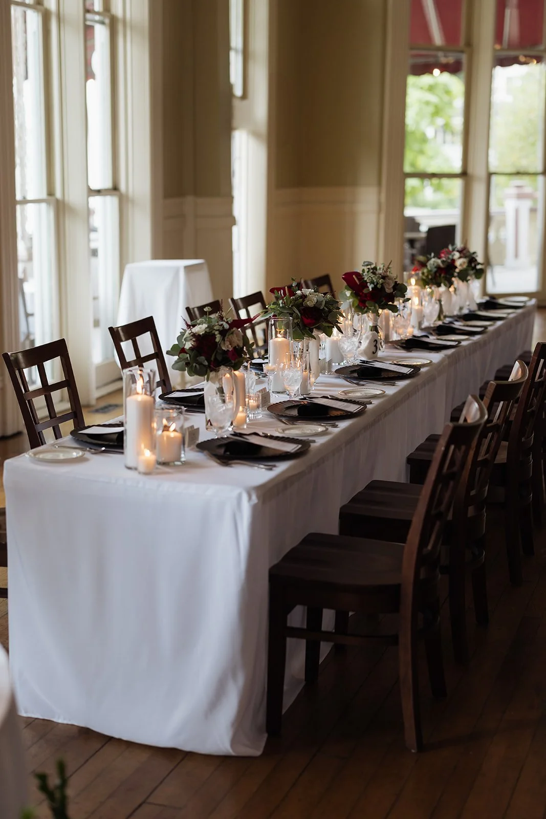 Elegant indoor wedding or event reception table set with white tablecloth, black napkins, glassware, candles, and floral centerpieces with red and white flowers and greenery. Wooden chairs surround the table, and natural light streams in from large w
