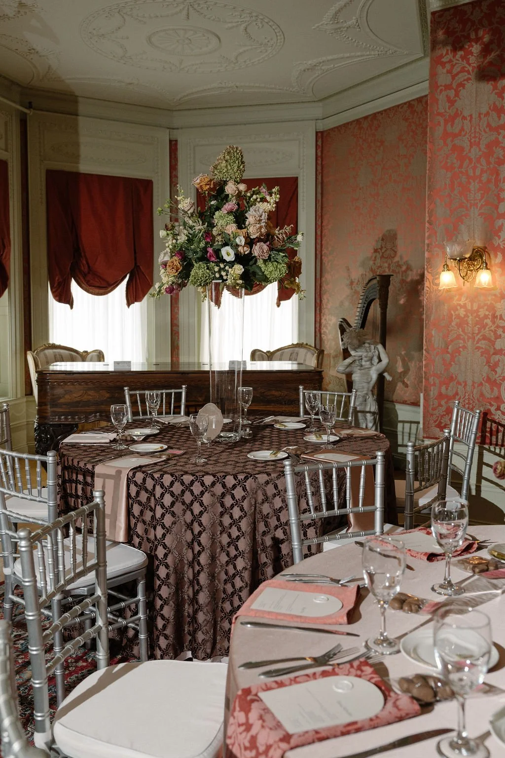 Elegant dining room with ornate white ceiling, pink patterned wallpaper, a large floral centerpiece on a table with a patterned tablecloth, and tables set with glassware, plates, and silverware.