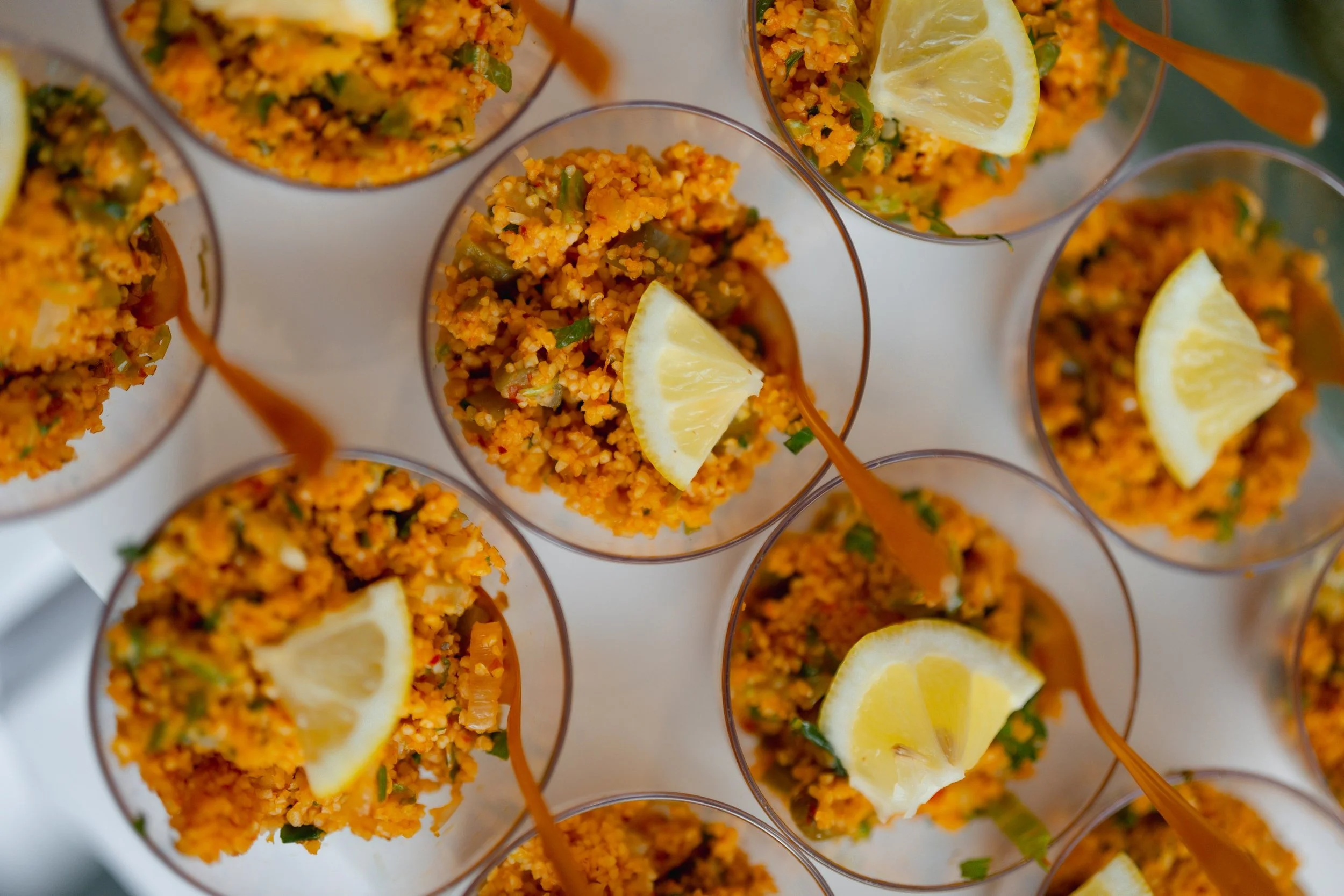 Close-up view of multiple servings of cooked couscous with herbs and vegetables in clear glasses, garnished with lemon wedges.