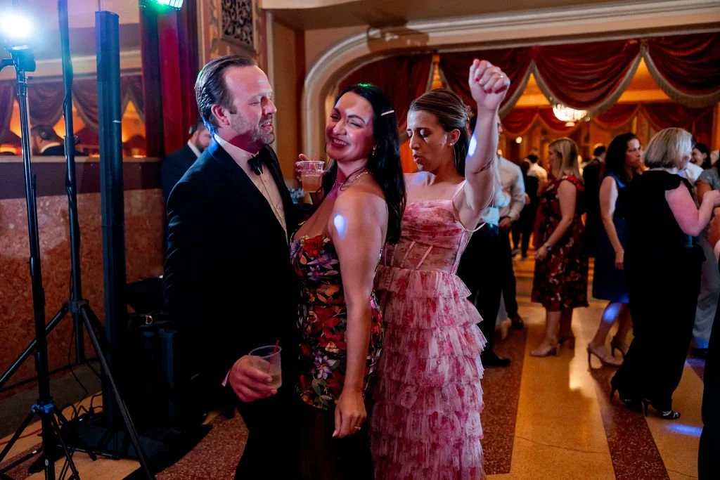 People dancing and socializing at a party in a decorated ballroom with red curtains and warm lighting.