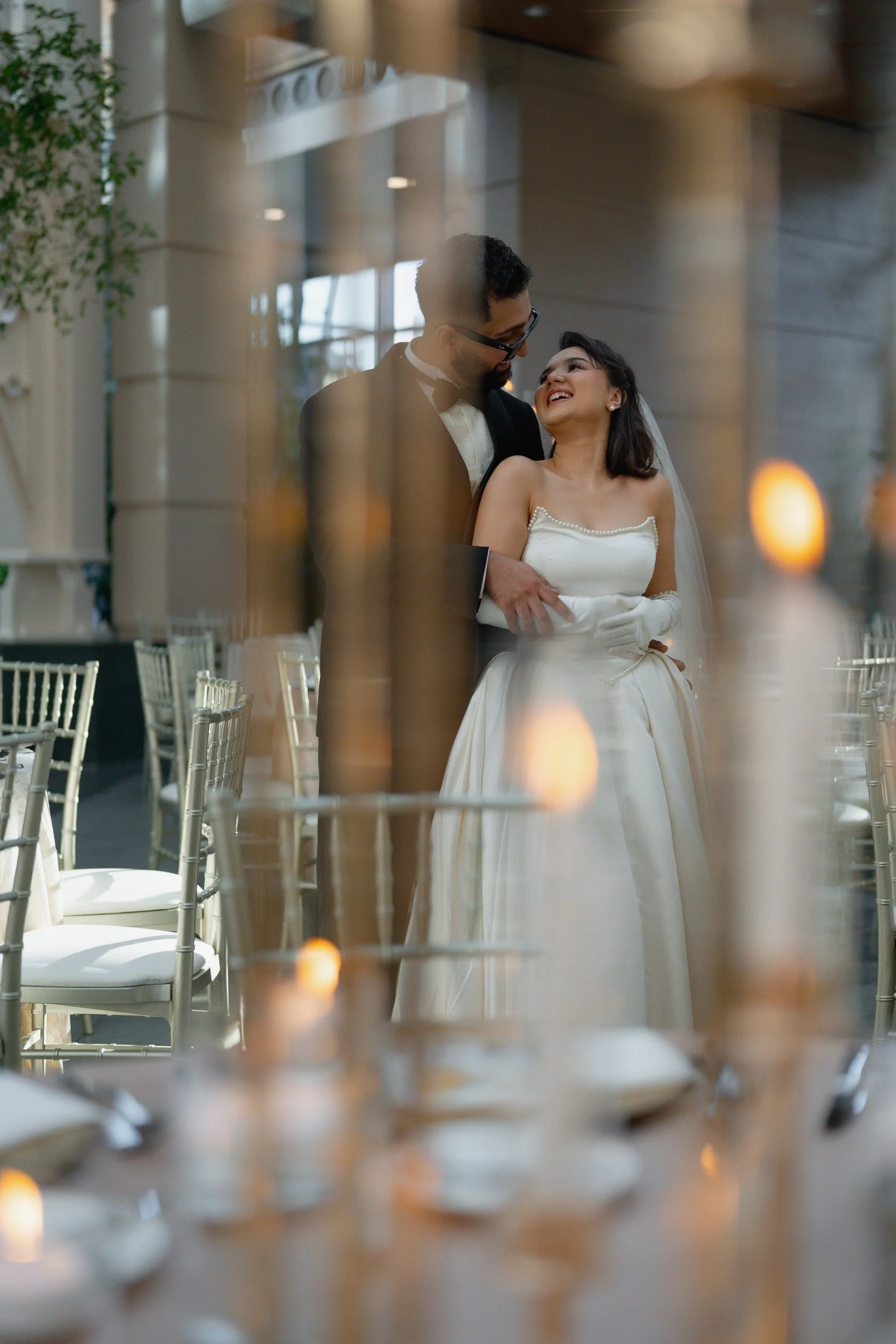 A newlywed couple sharing a happy moment, smiling at each other, at their wedding reception. The bride is wearing a strapless wedding gown with pearl details, and the groom is dressed in a black tuxedo with a bow tie. The photo is taken through a ref