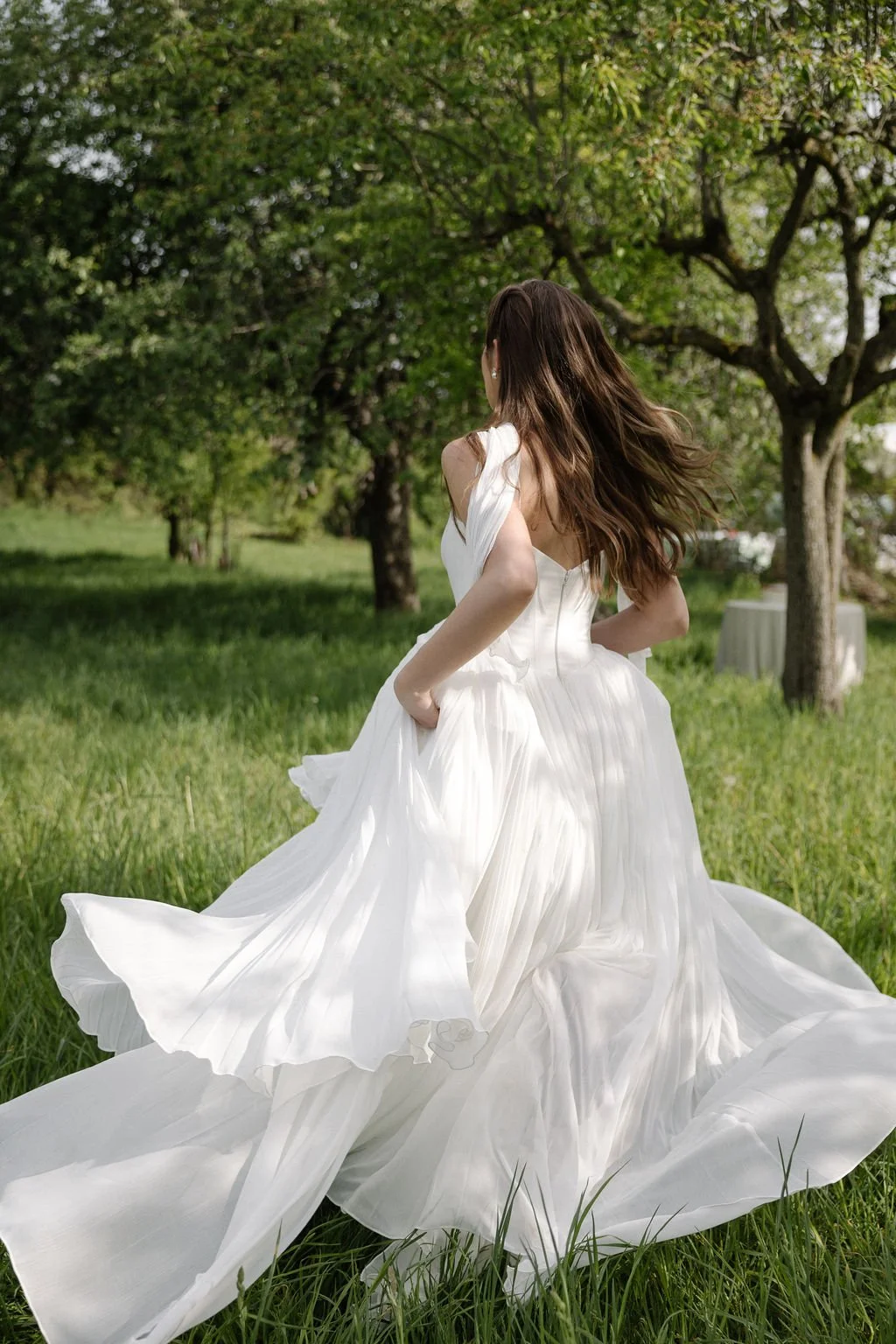 A woman in a flowing white dress standing in a grassy field with trees in the background.