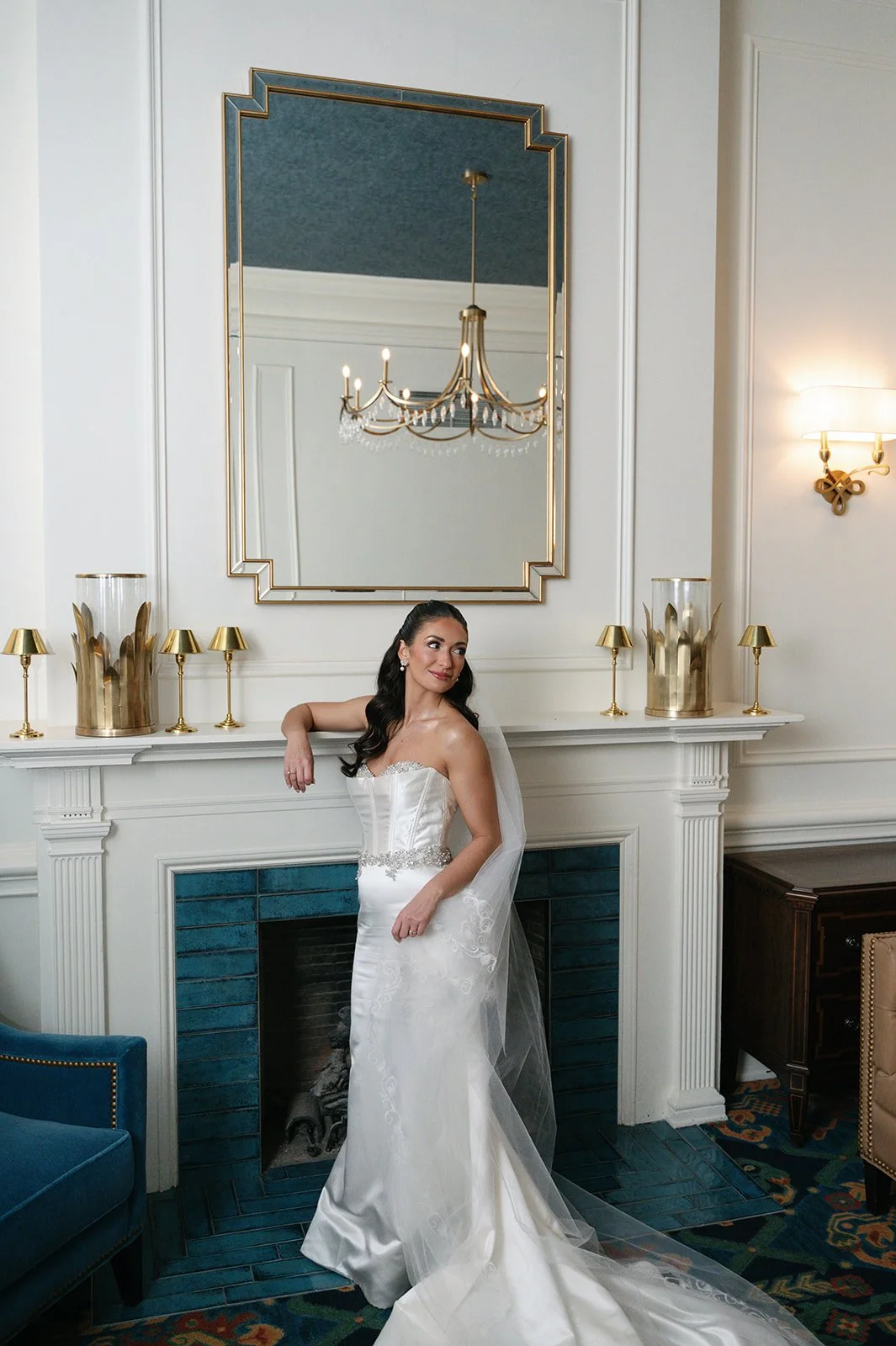 A woman in a white wedding dress standing in front of a fireplace with a large mirror above it, surrounded by gold candle holders and decorative items, in a elegantly decorated room.