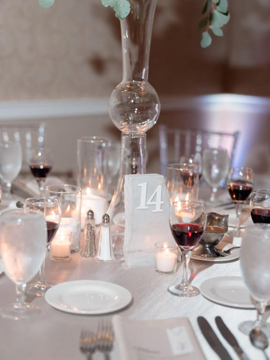 Elegant table setting with wine glasses, tealight candles, a stone table number with the number 14, a tall glass centerpiece, and a white tablecloth.