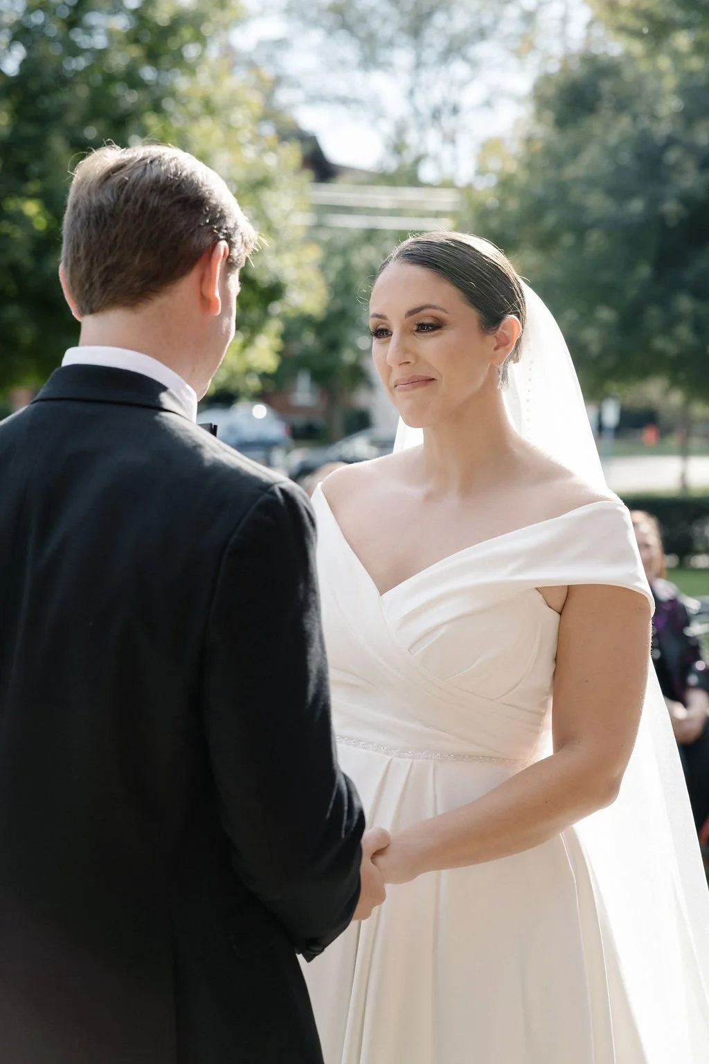 A bride and groom holding hands during their wedding ceremony outdoors with trees and sunlight in the background.